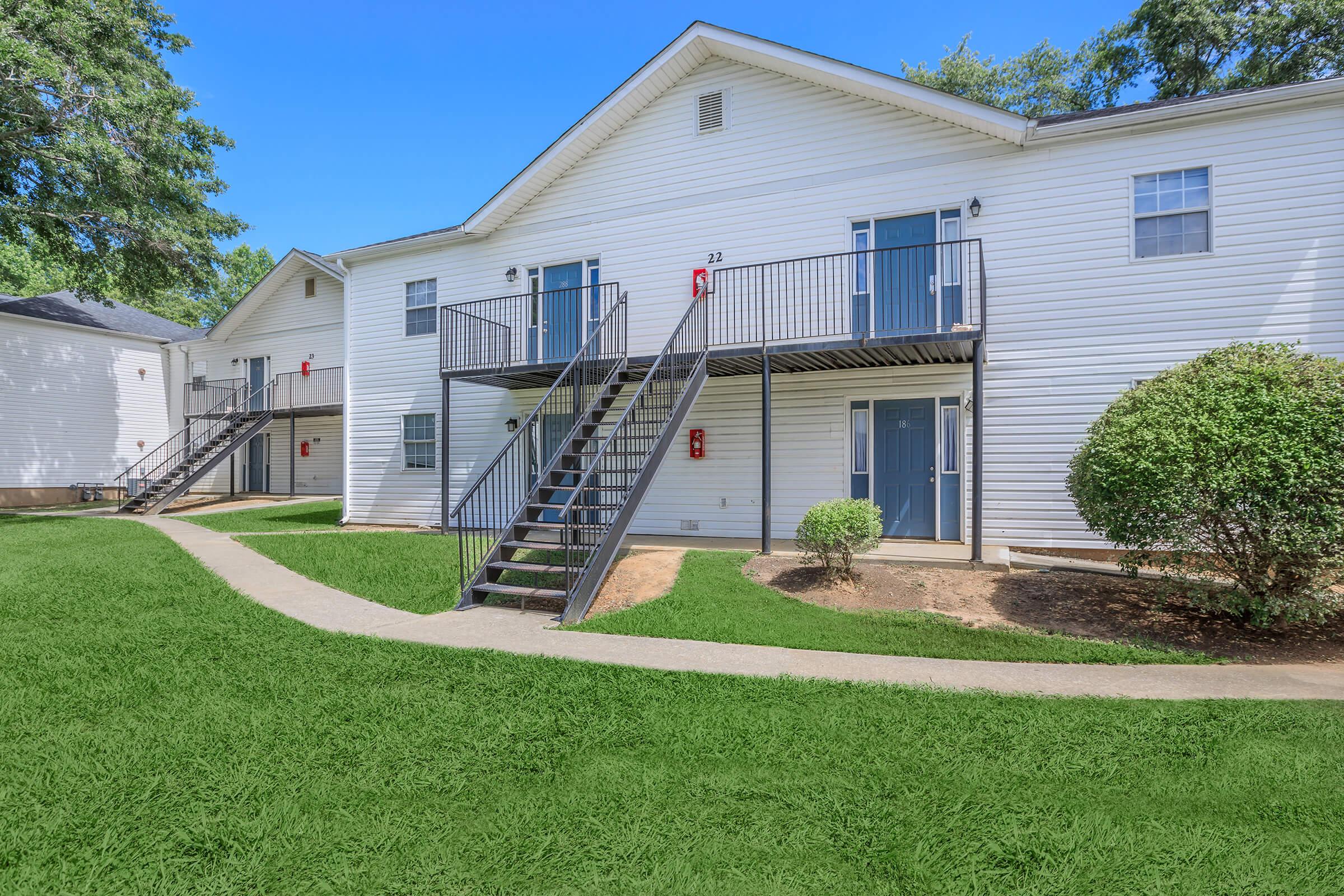A view of a two-story apartment complex with white siding. There are multiple doors on the upper and lower levels, and a set of stairs leads to the second floor. The area is well-maintained with green grass and a curved pathway. Trees are visible in the background, providing a pleasant environment.