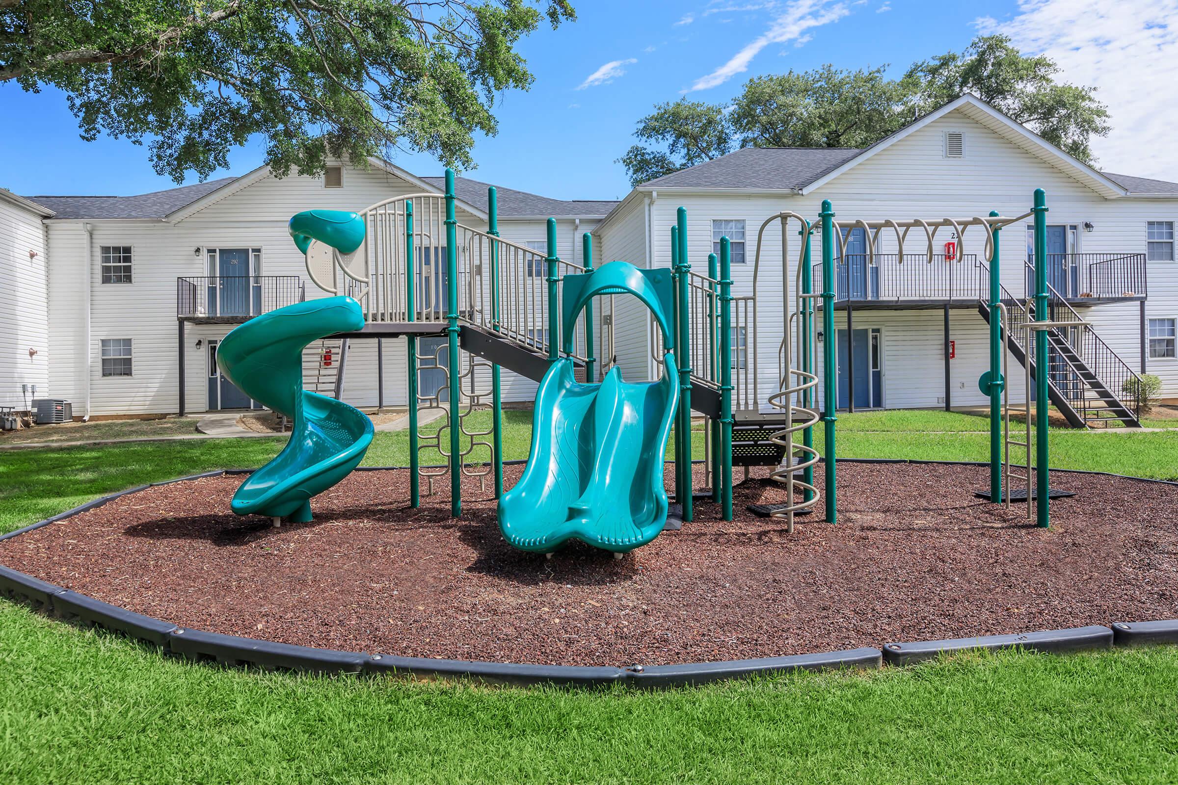 Colorful playground equipment featuring a green spiral slide and two side-by-side slides, surrounded by a soft mulch surface. In the background, there are white apartment buildings with balconies and trees providing shade under a clear blue sky.