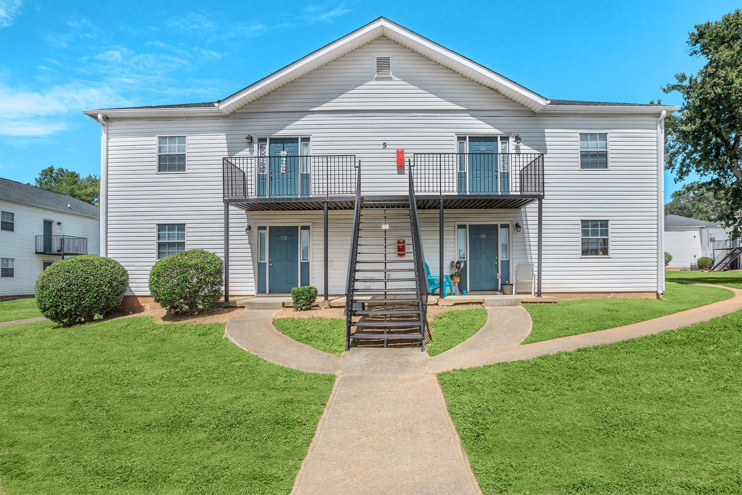 Two-story apartment building with a white exterior, featuring three staircases leading to separate entrances. The lawn is well-maintained with green grass and small bushes, and a clear blue sky is visible in the background.