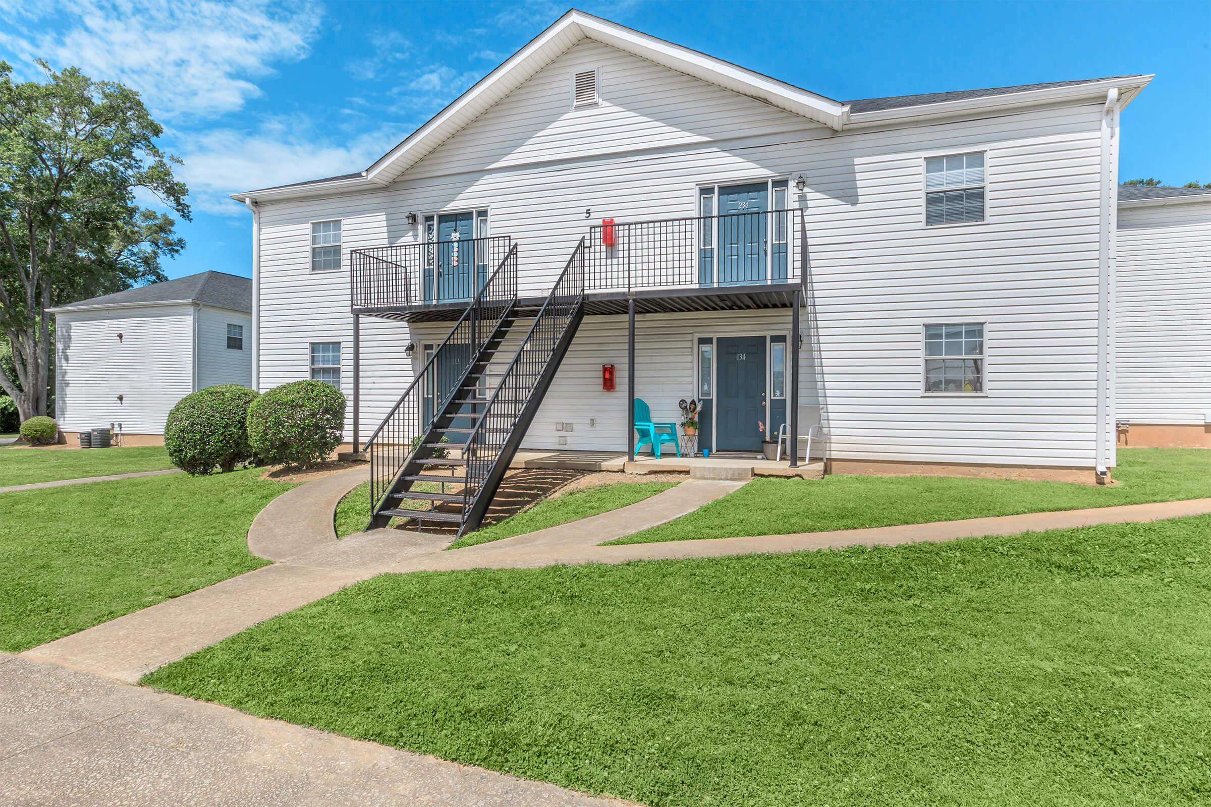 Two-story apartment building with white siding, featuring a staircase leading to the upper units. Each unit has a balcony with chairs and a red fire extinguisher visible. The area includes well-kept lawns and a concrete walkway. Clear blue sky and some trees are in the background.
