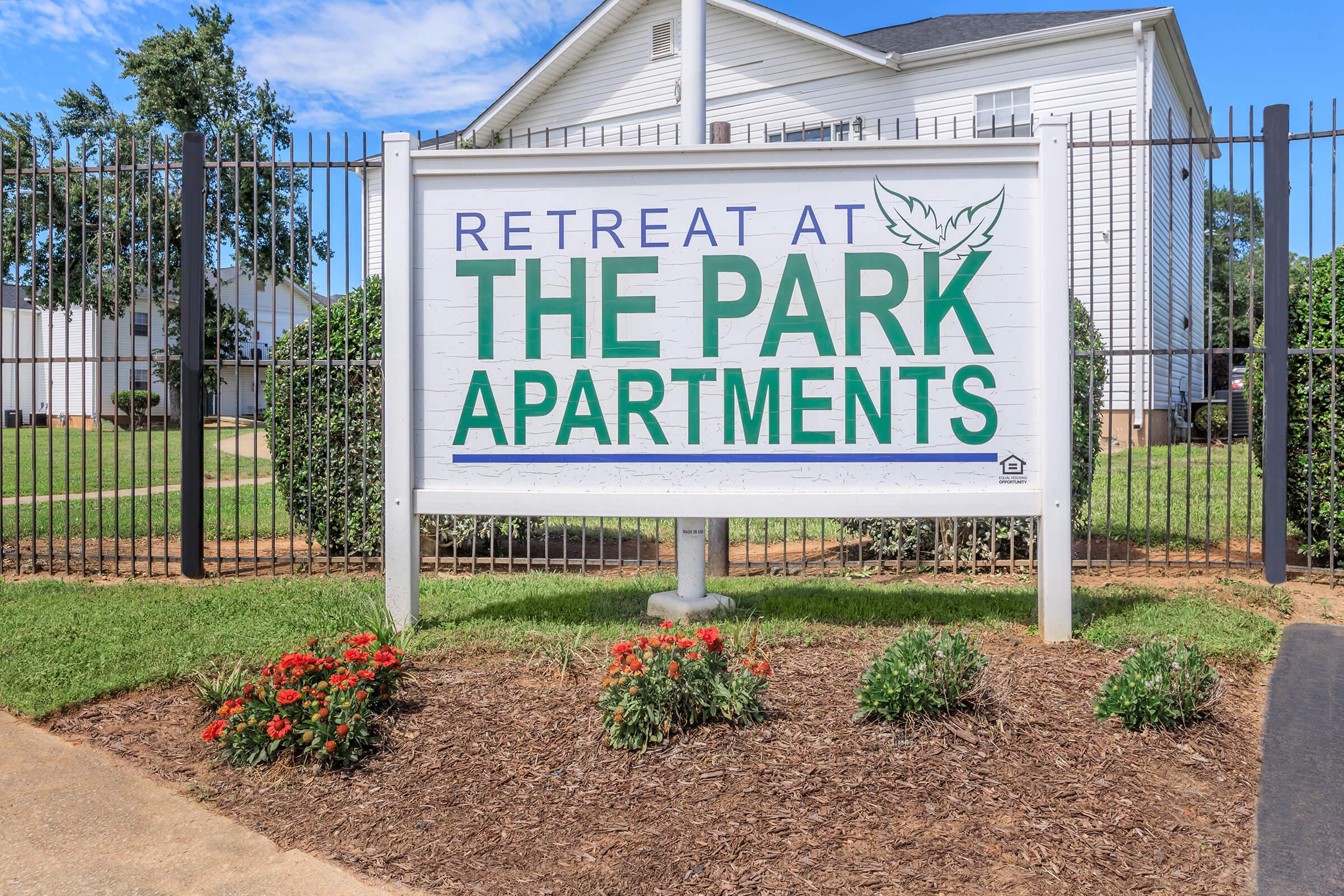 Sign for "Retreat at The Park Apartments" in front of a well-maintained property, featuring green grass and flower beds. A fence and a white building are visible in the background, indicating a residential complex.