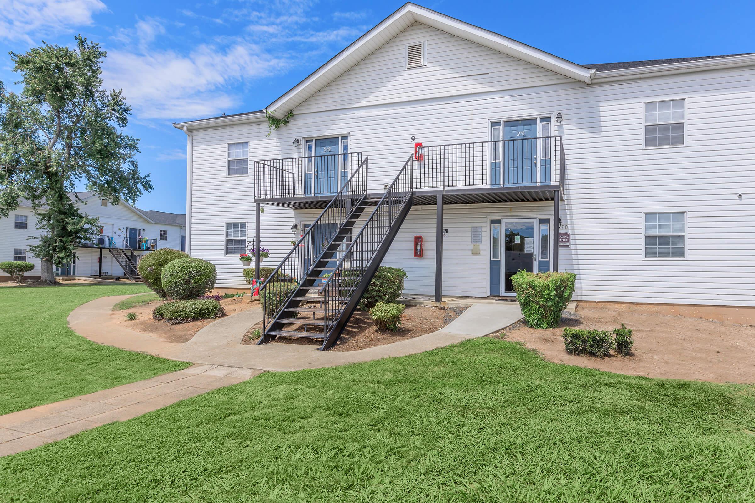 Two-story apartment building with white siding. The front features a set of black metal stairs leading to two entrance doors on the upper level, with balconies. The lawn is green, and there are neatly trimmed bushes and a paved walkway leading up to the building. Clear blue sky in the background.