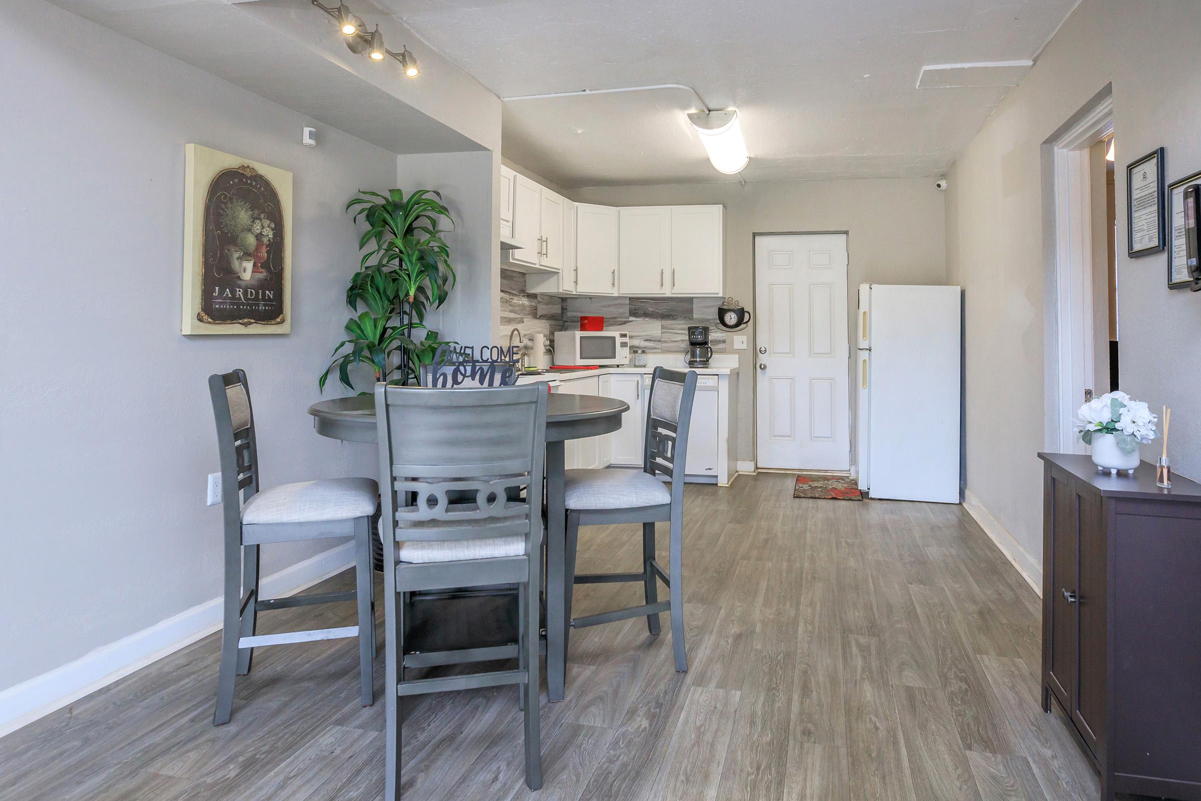 Modern kitchen and dining area featuring a round wooden table with four chairs, a potted plant, white cabinets, and stainless steel appliances. The decor includes a framed artwork on the wall and a door leading to an adjacent room. The space has light-colored walls and vinyl flooring.