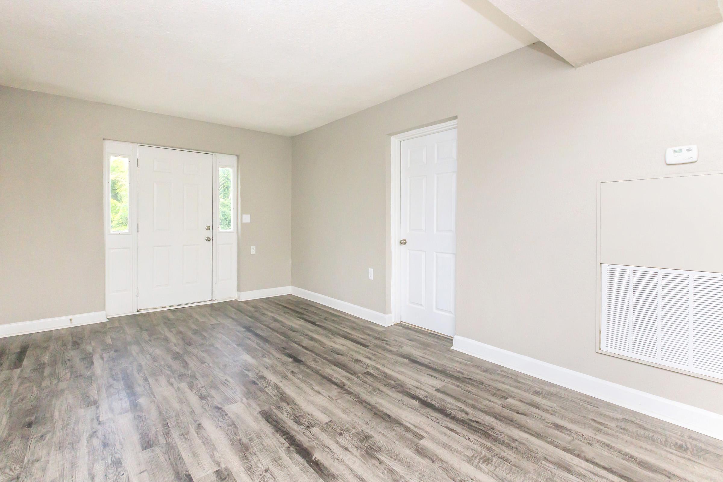 A spacious, empty room with light gray walls and new laminate flooring. There are two closed doors, one leading outside and another into a separate room. A wall vent is visible on the right side. Natural light enters through the front door, creating a bright atmosphere.