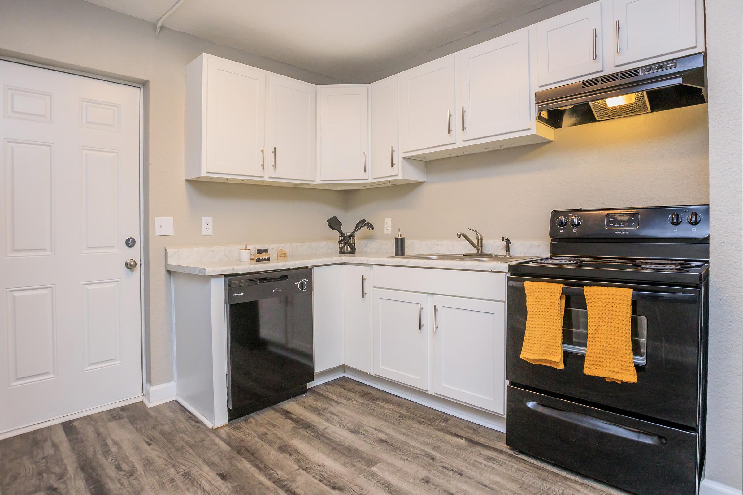 Modern kitchen featuring white cabinets, a black stove, and a dishwasher. The countertop is light-colored, with a sink and faucet, and a black kitchen utensil holder. There are two orange towels hanging from the oven handle, and a door is visible on the left side of the image. The flooring is a dark wood look.