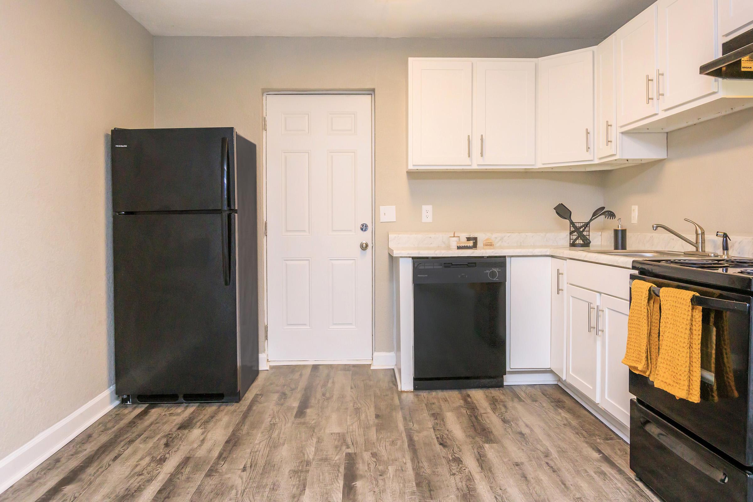 A modern kitchen featuring a black refrigerator, white cabinets, a countertop, a black dishwasher, and a black stove. The floor is covered in gray wood-like tiles. A door leads to an exterior space, and yellow towels hang from the stove. The walls are painted in a light beige color.