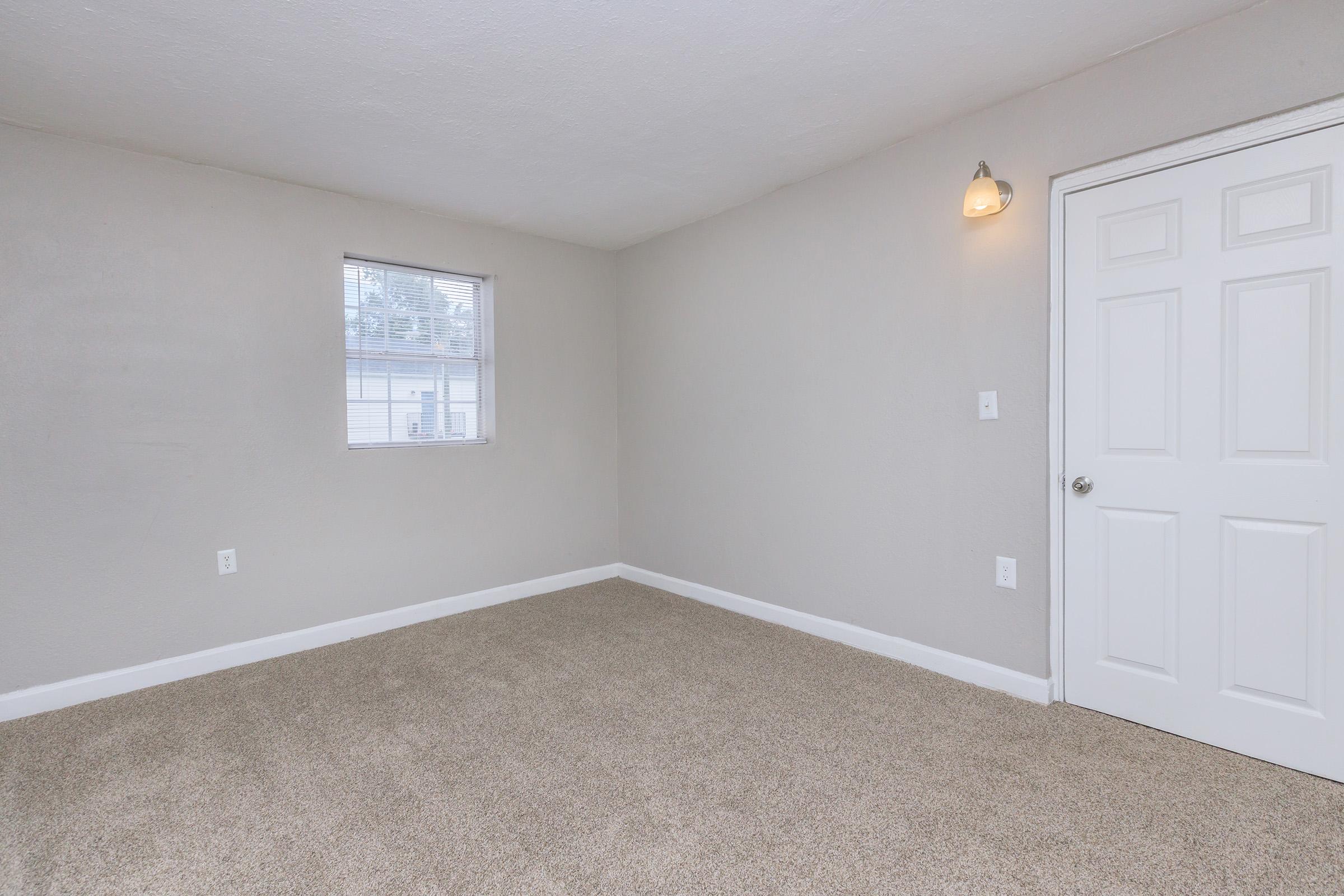 A small, empty room with light gray walls and a window on one side. The floor is covered in beige carpet, creating a cozy atmosphere. There's a light fixture on the wall and a white door leading to another area. The overall appearance is clean and minimalistic, ideal for various uses.