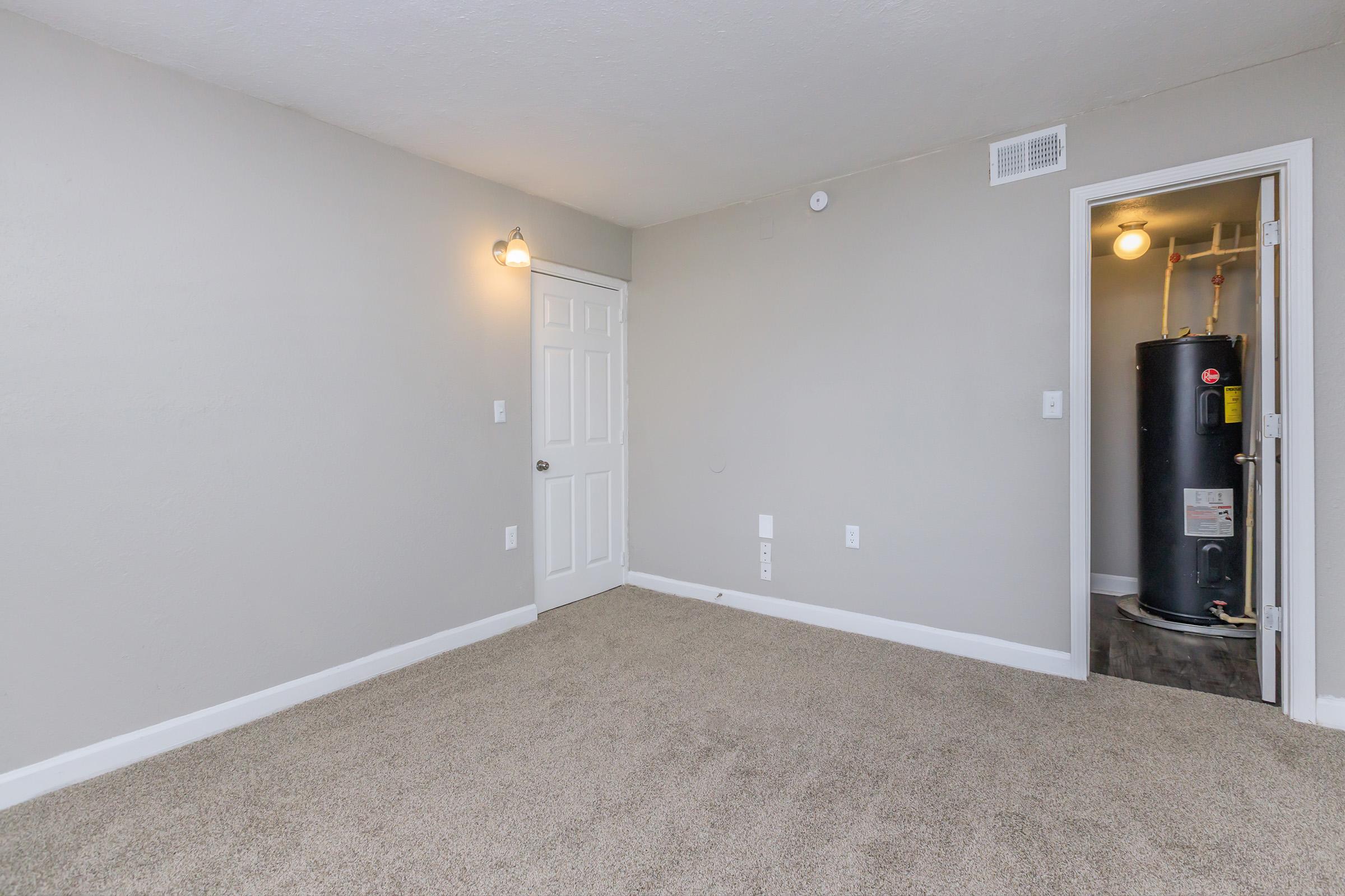 A small, empty room with light gray walls and carpeted flooring. There is a door on the left side, and a water heater visible in a closet to the right. Two wall-mounted light fixtures provide illumination. The overall space appears clean and well-maintained.