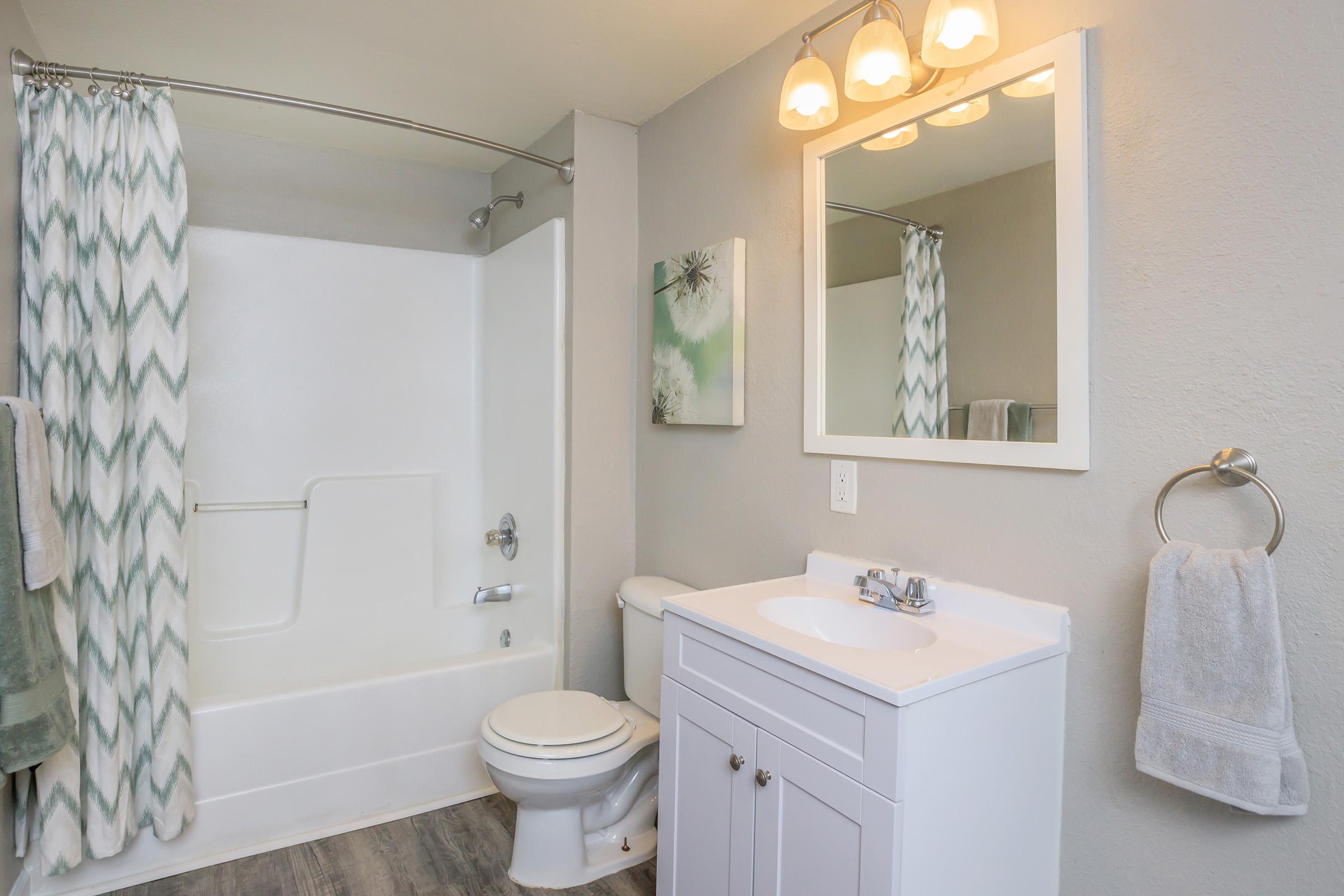 A modern bathroom featuring a bathtub with a shower curtain, a white vanity with a sink, and a mirror above it. Soft green and gray tones create a calming atmosphere. There is an abstract wall art piece and neatly arranged towels beside the sink. Bright lighting enhances the clean and inviting look.