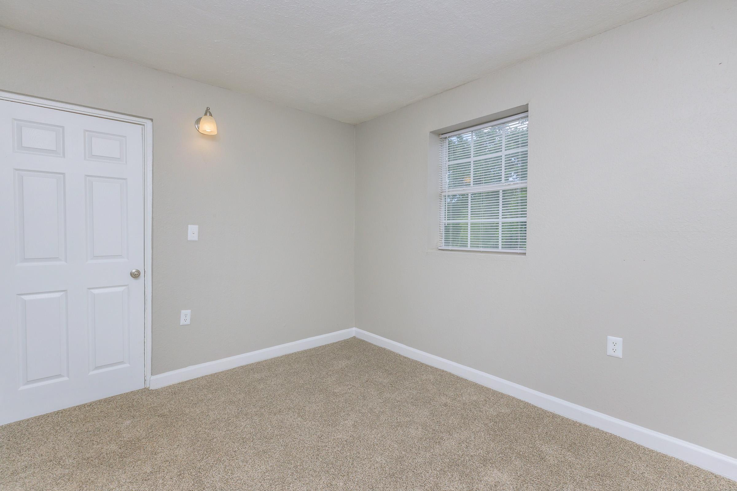 Empty room with light beige walls, a window with white blinds, and carpeted flooring. There is a single light fixture on the wall and a closed white door. The space is bright and minimalistic, creating a neutral atmosphere.