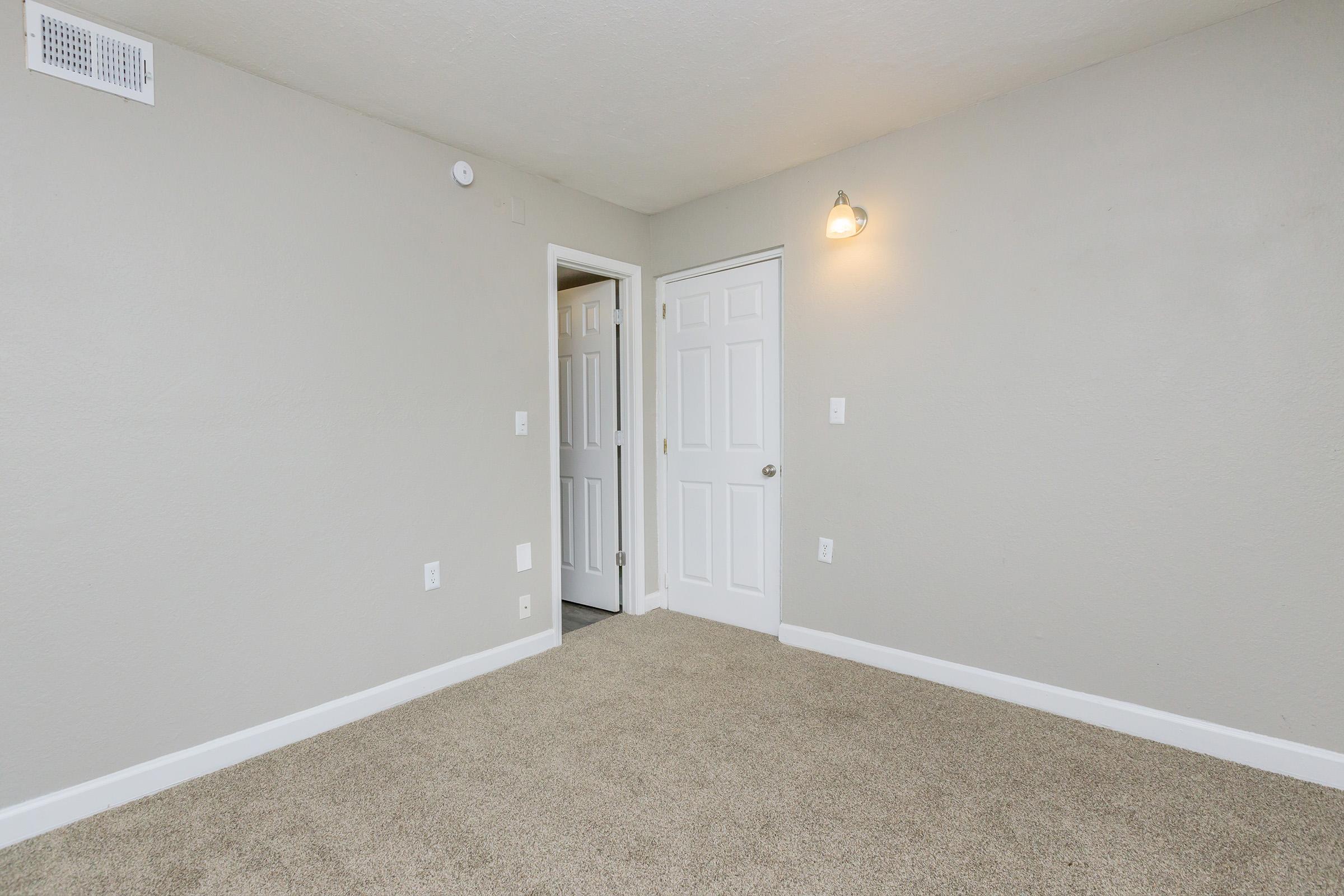 A light gray room with beige carpet flooring. There are two white doors: one partially open leading to another space. A wall-mounted light fixture illuminates the room, which has minimal decor and a clean, uncluttered appearance.