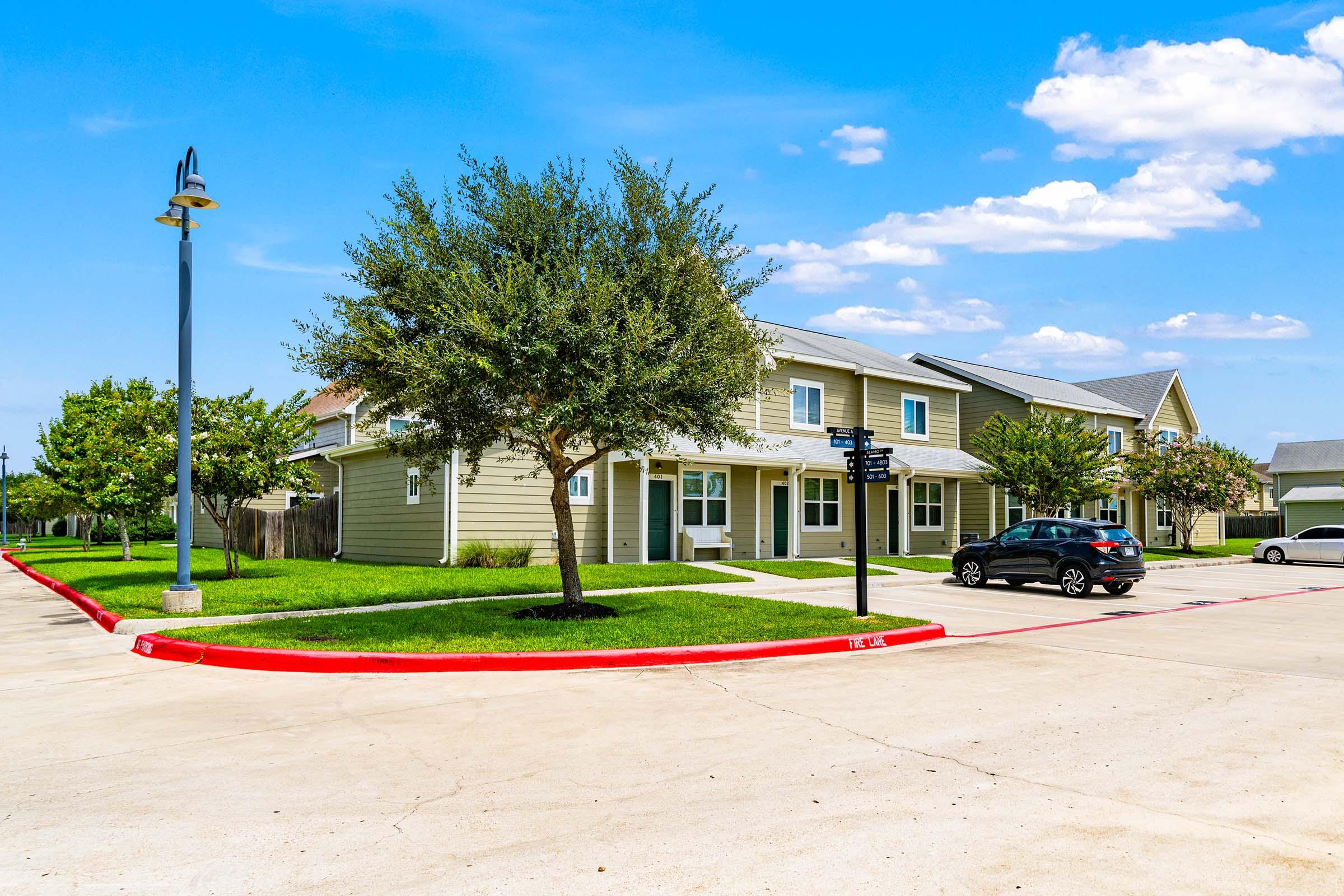 A residential area featuring two-story houses, surrounded by lush green lawns and trees. A black car is parked along a red-bordered parking lot, with lampposts and blue sky with scattered clouds in the background.