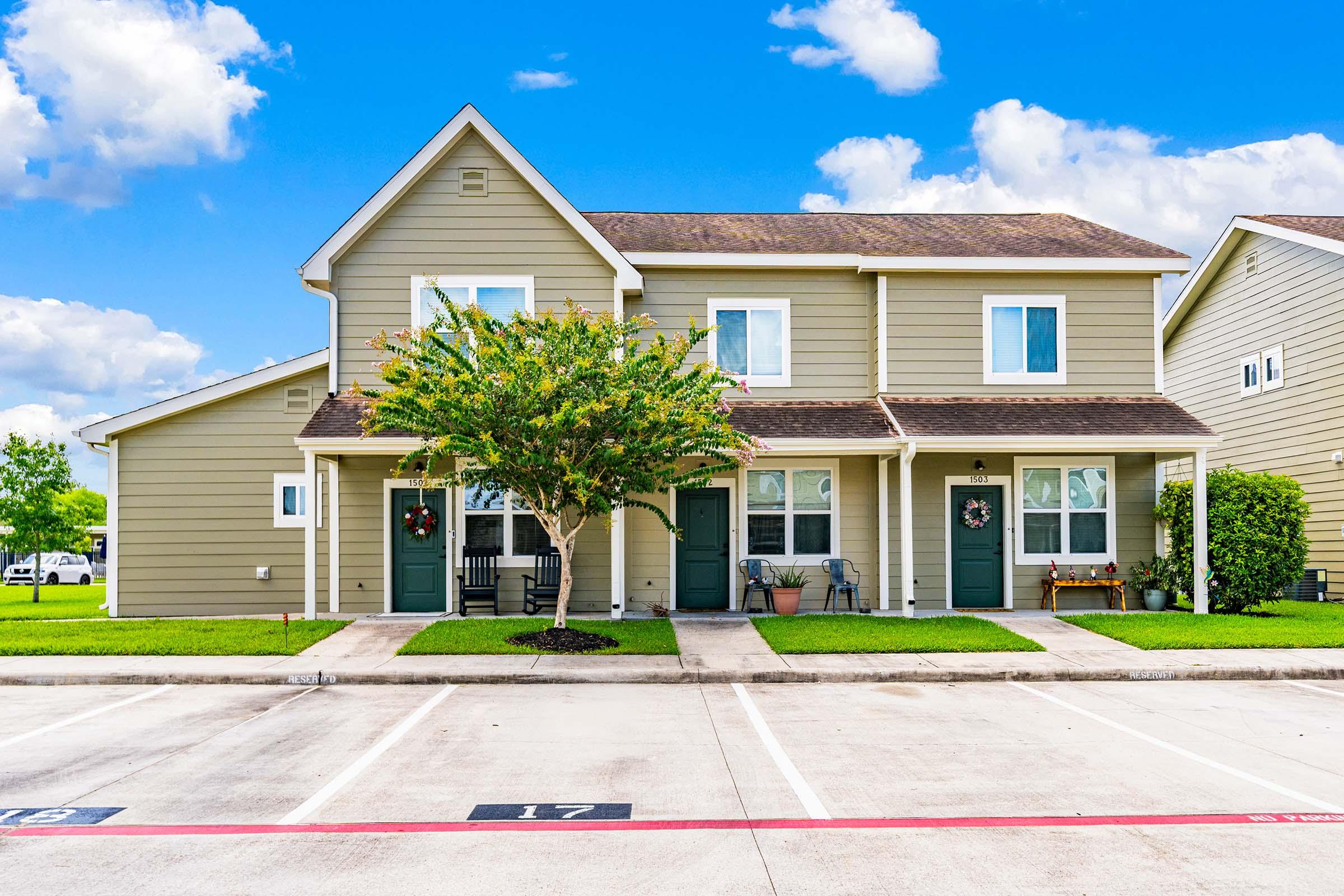 Two-story duplex with light green siding, featuring a front porch area, neatly trimmed grass, and a small tree in front. The building has decorative wreaths on the doors and is surrounded by parking spaces under a blue sky with fluffy white clouds.