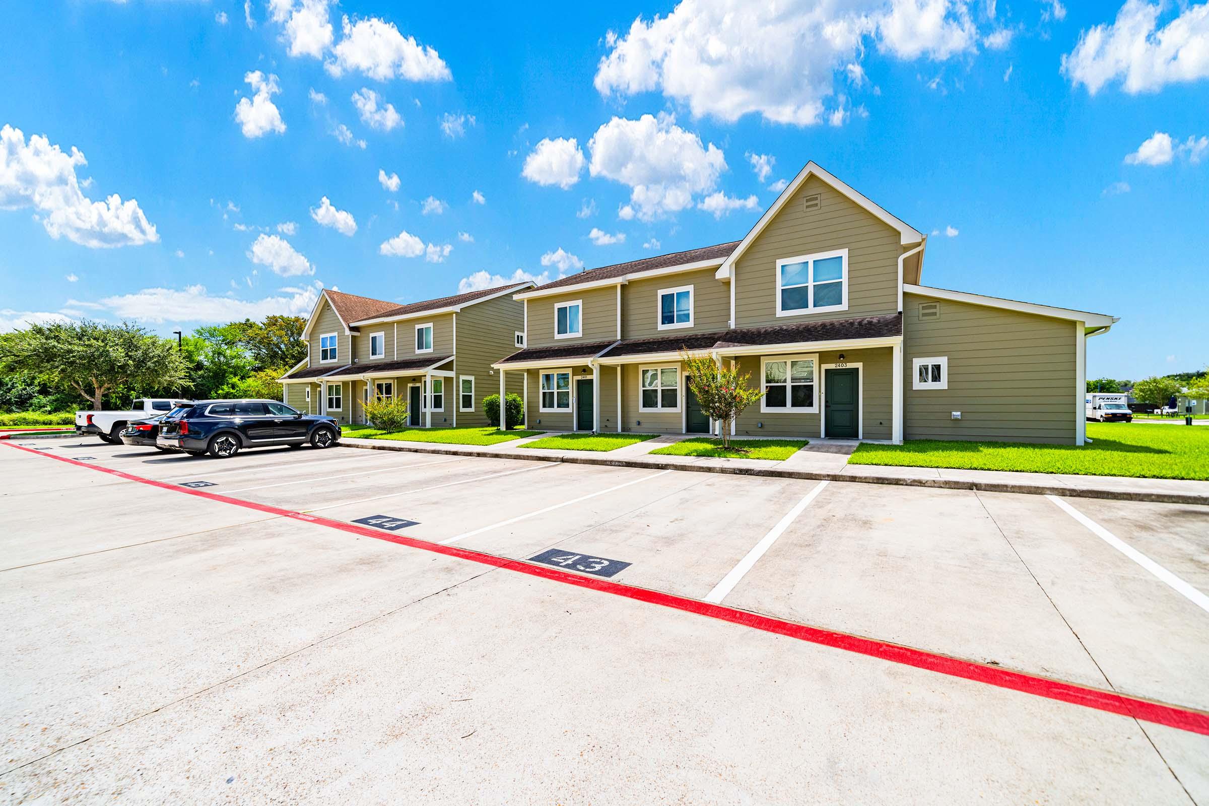 A clean and well-maintained parking lot in front of two-story residential buildings. The buildings are painted in light colors with well-manicured lawns. Several parked cars are visible along the designated parking spaces, and the sky is bright blue with fluffy white clouds.