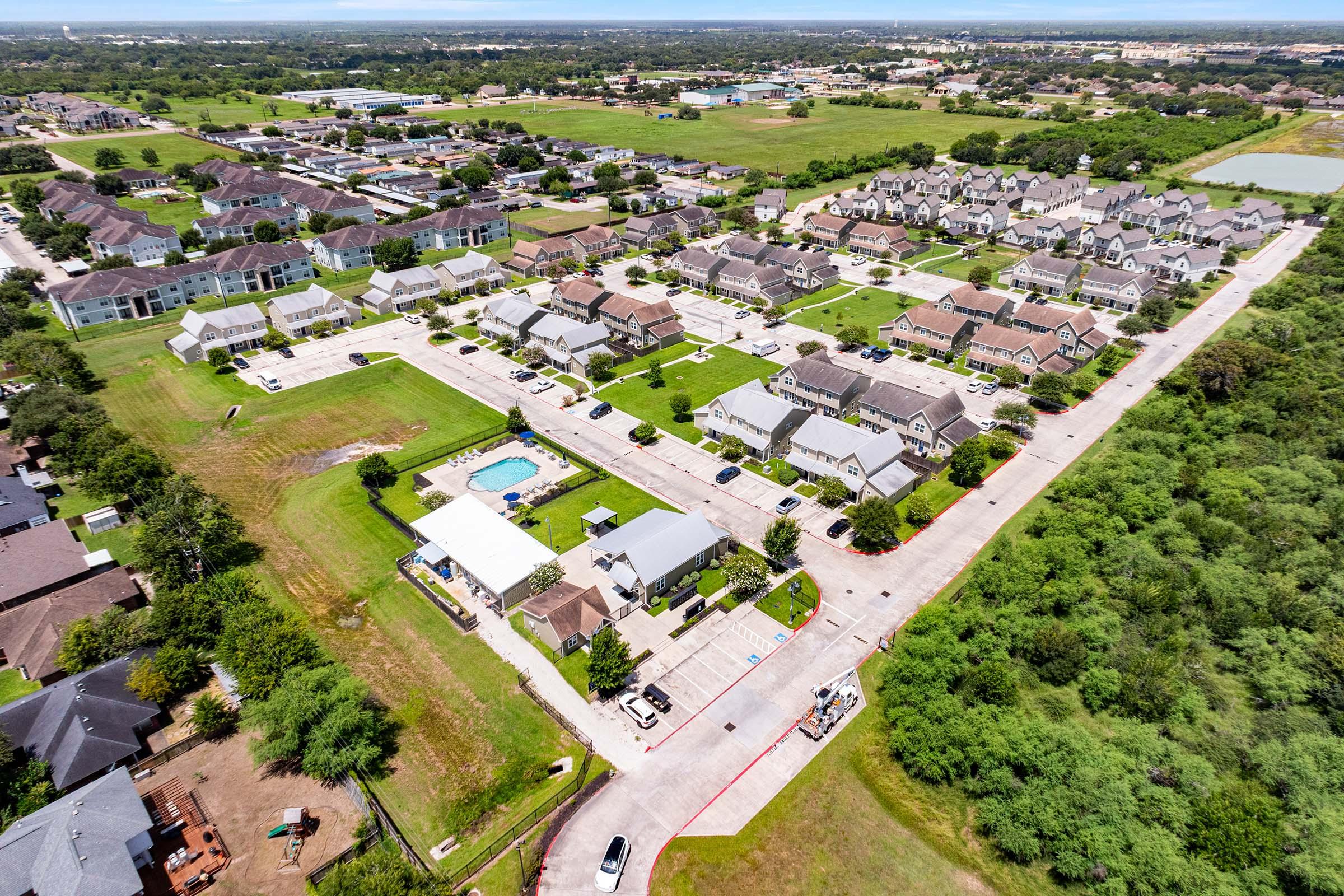Aerial view of a residential neighborhood featuring rows of houses, green lawns, and a community pool. The area is surrounded by lush greenery and open fields, with nearby commercial buildings visible in the distance. The layout includes paved roads and parking spaces, indicating a suburban community.