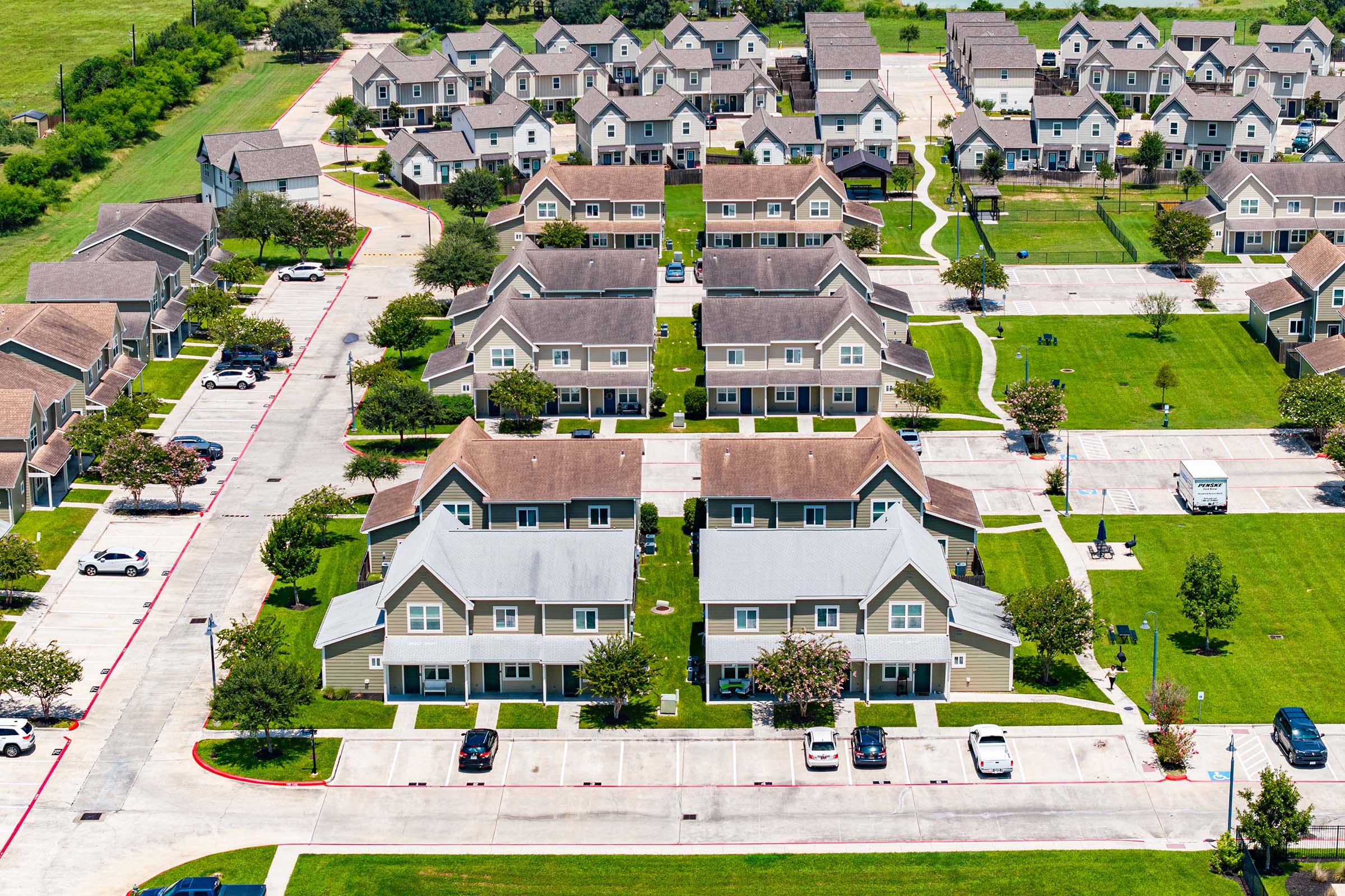 Aerial view of a suburban neighborhood featuring multiple two-story houses with pitched roofs, front lawns, and driveways. The area is well-maintained with green grass and trees, and parking spaces are visible along the roads. The layout is organized in a grid pattern, showcasing a modern community design.