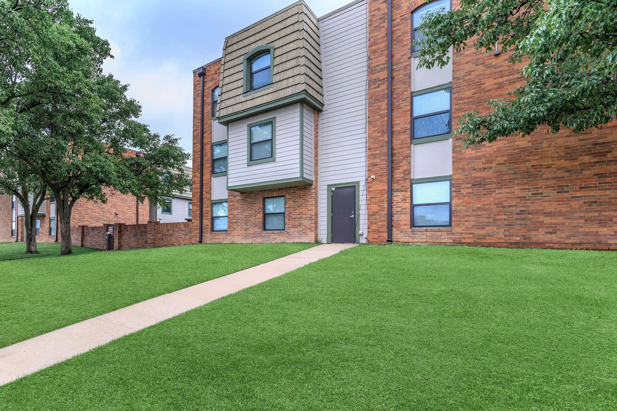 A modern apartment building featuring a mixture of brick and siding. The building has a unique, sloped roof on one section and several windows. A well-maintained green lawn is in front, with a concrete pathway leading to the entrance. Trees are visible on either side of the building.