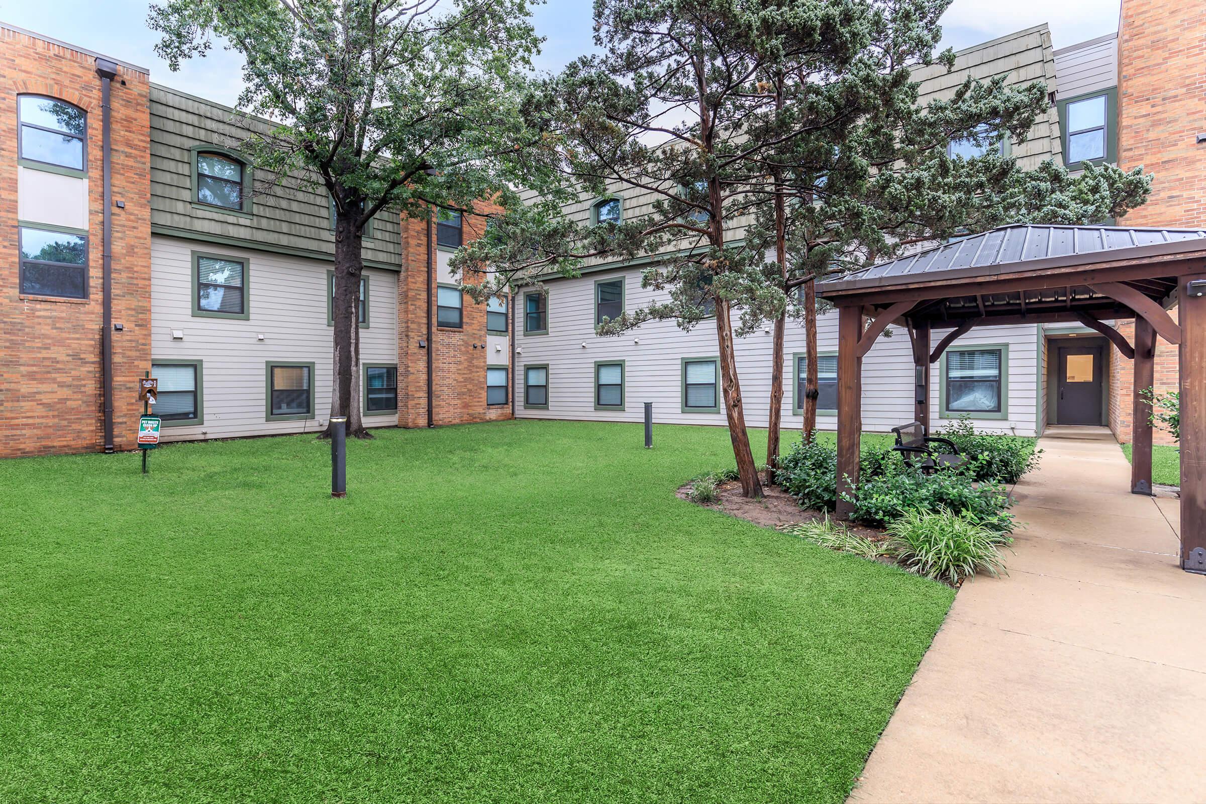 A landscaped courtyard featuring well-maintained green grass, trees, and shrubs, surrounded by a building with a mix of brick and siding exteriors. A covered gazebo is visible in one corner, providing a seating area. The overall atmosphere is inviting and serene, ideal for outdoor relaxation.