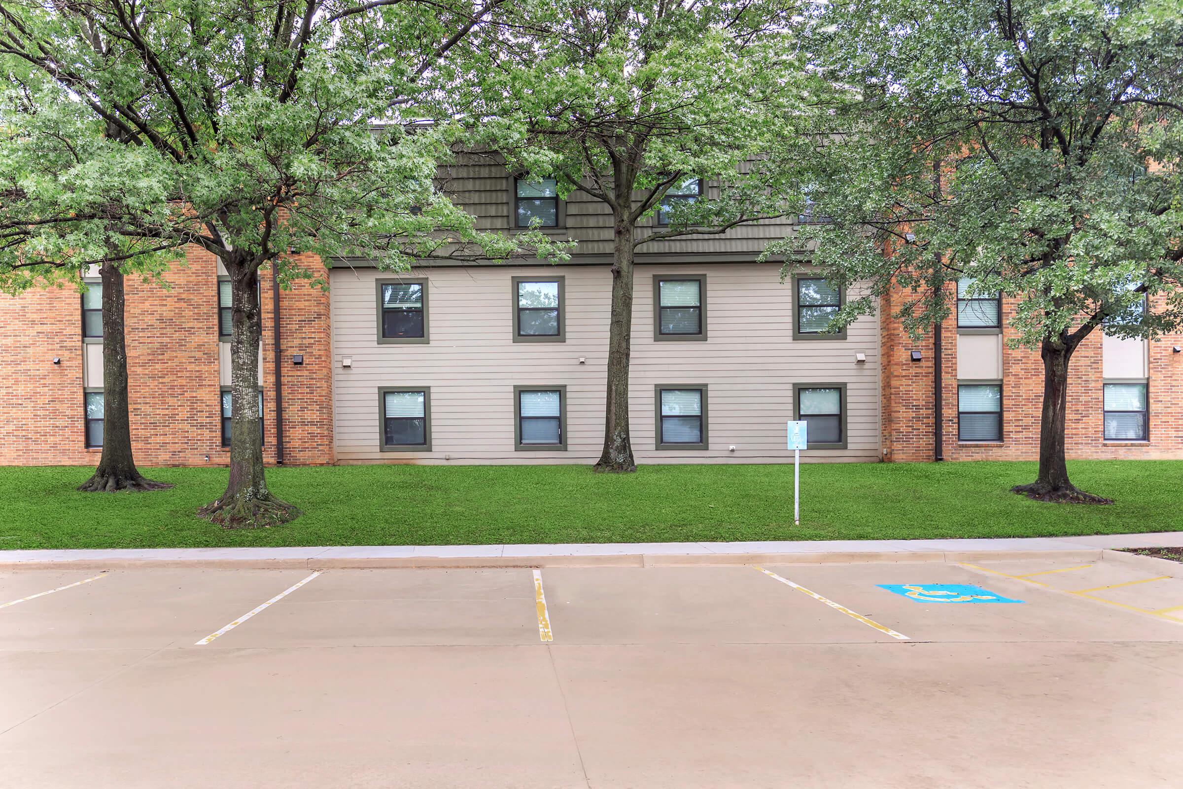 A view of a multi-story building with a mix of brick and siding exterior. Two trees are planted in front of the building, and there is a grassy area as well as a parking lot with marked spaces, including a spot for disabled parking. The sky is overcast, indicating a cloudy day.