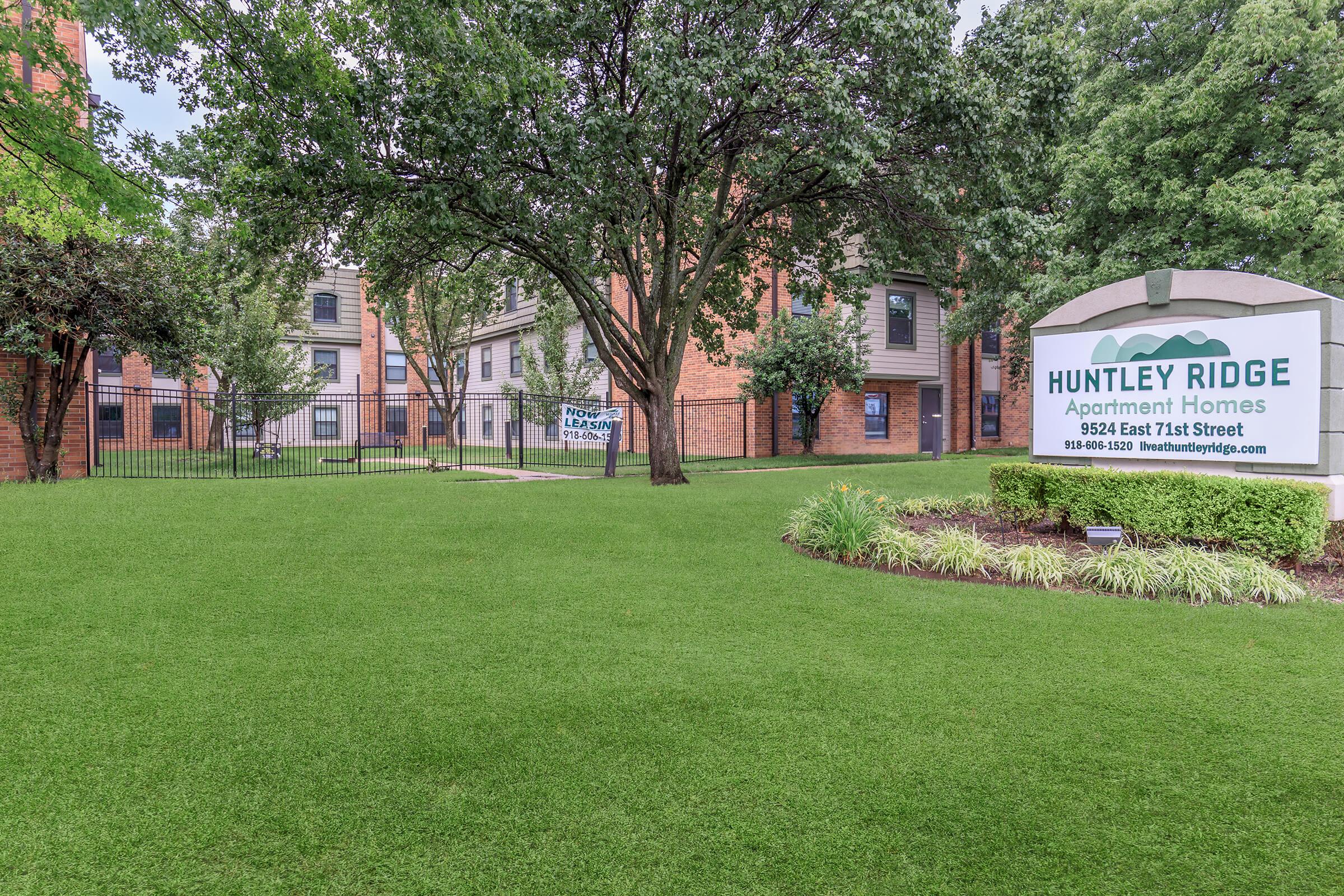 Sign for Huntley Ridge Apartment Homes in a well-maintained grassy area, surrounded by trees and a glimpse of apartment buildings in the background. The scene conveys a welcoming atmosphere for potential residents.
