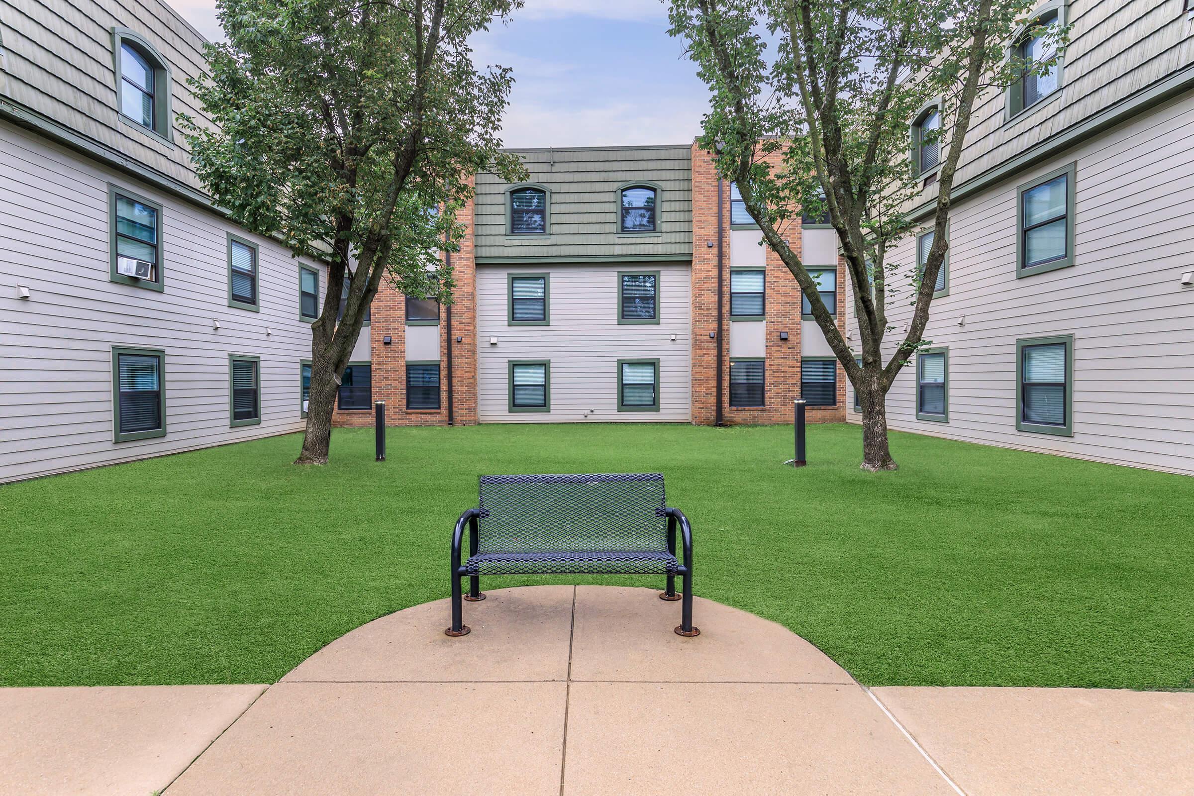 A small courtyard surrounded by residential buildings with two trees and a green lawn. In the center, there is a blue metal bench on a concrete path, providing a place to sit and enjoy the outdoor space. The buildings have a modern design with multiple windows.