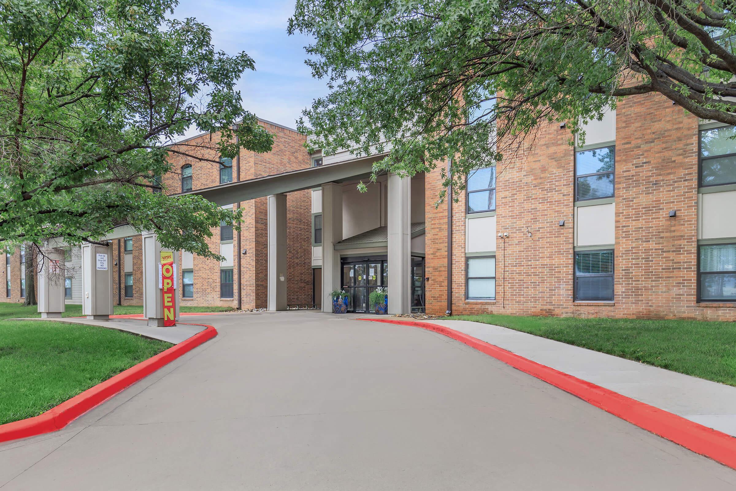 A view of a brick building with a welcoming entrance. A large "OPEN" sign is prominently displayed at the front. The path leading to the entrance is lined with green grass and trees, creating a pleasant environment. The sky is blue with some clouds, adding to the inviting atmosphere.