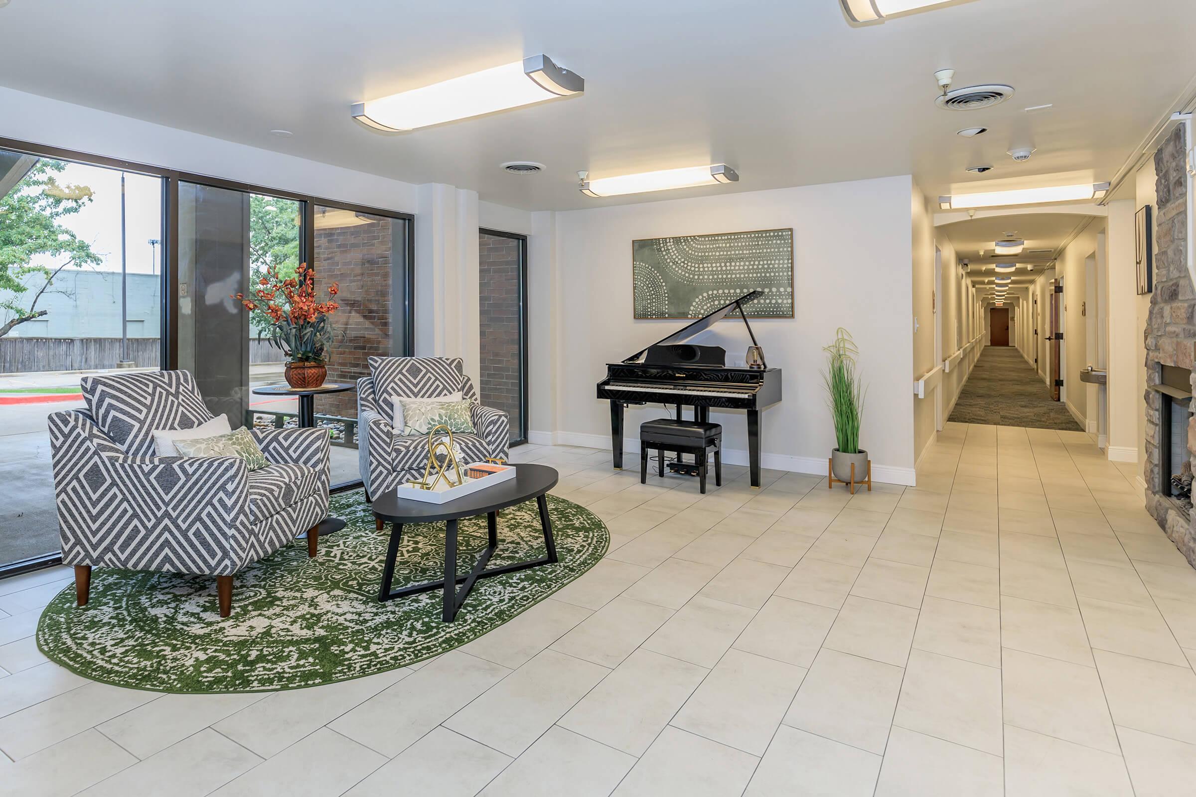 A bright hallway featuring a black piano, two patterned armchairs, and a small table. Decorative plants and a wall art piece enhance the ambiance. Natural light comes from large windows, and a long corridor extends in the background, leading to additional spaces.
