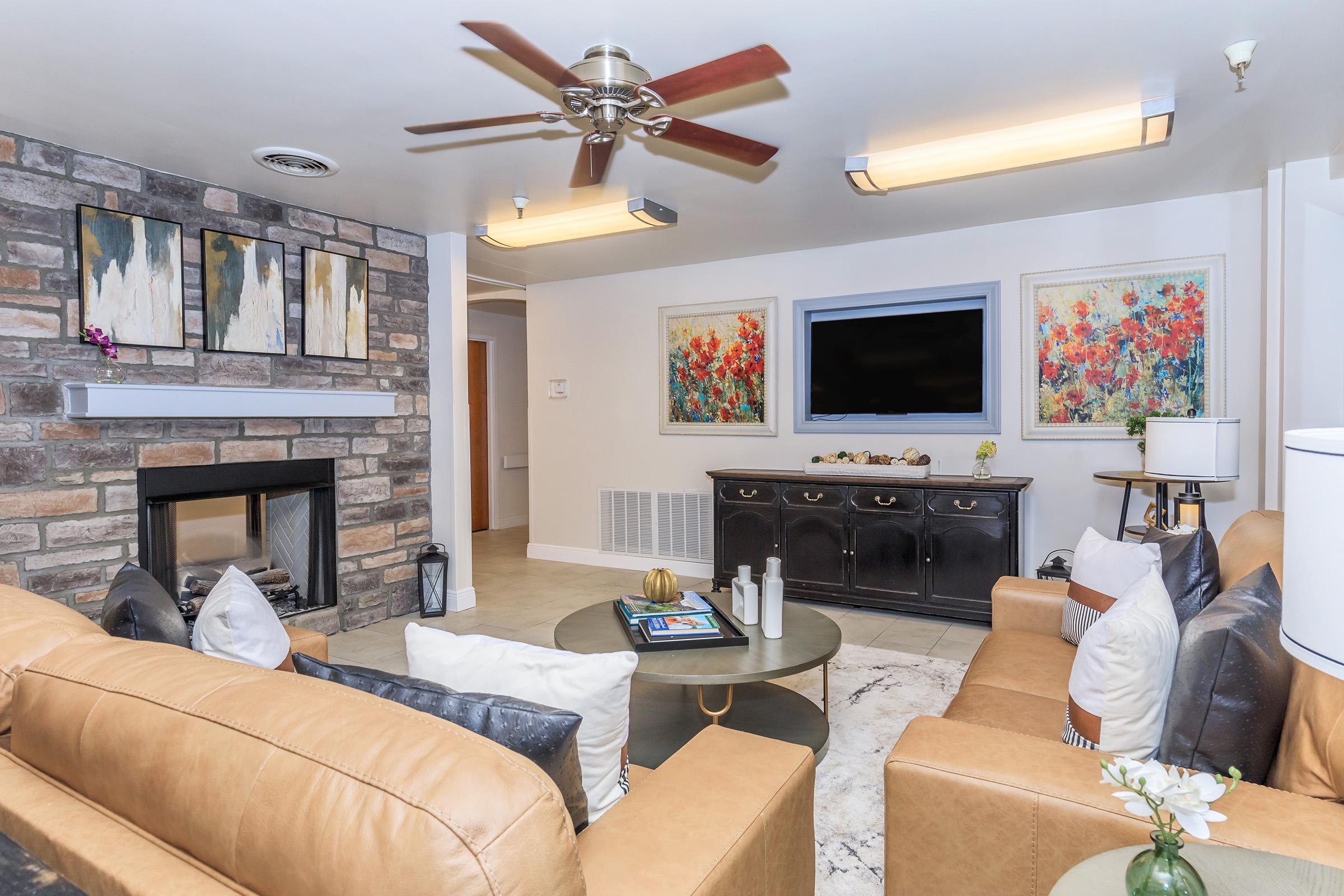 A cozy living room with a stone fireplace, two tan leather sofas, and a round coffee table. The wall features abstract artwork in vibrant colors, and a flat-screen TV is mounted above the fireplace. Plants and decorative items add a homey touch to the space.