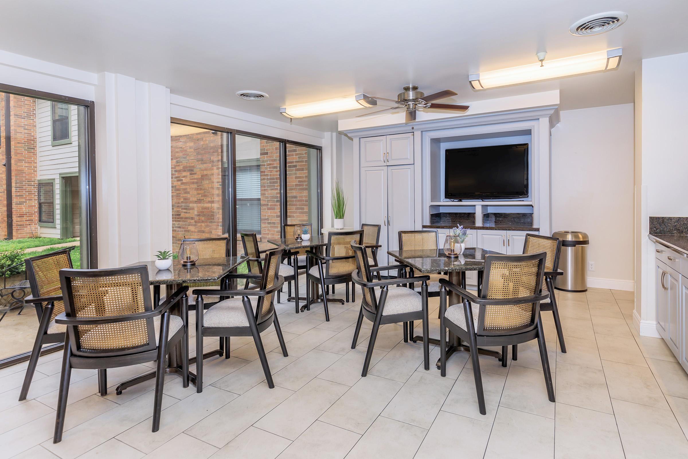 A bright, modern dining area featuring several tables with chairs, large windows allowing natural light, and a kitchenette in the background. There is a television mounted on the wall, indoor plants for decoration, and a clean, contemporary design that creates an inviting atmosphere.