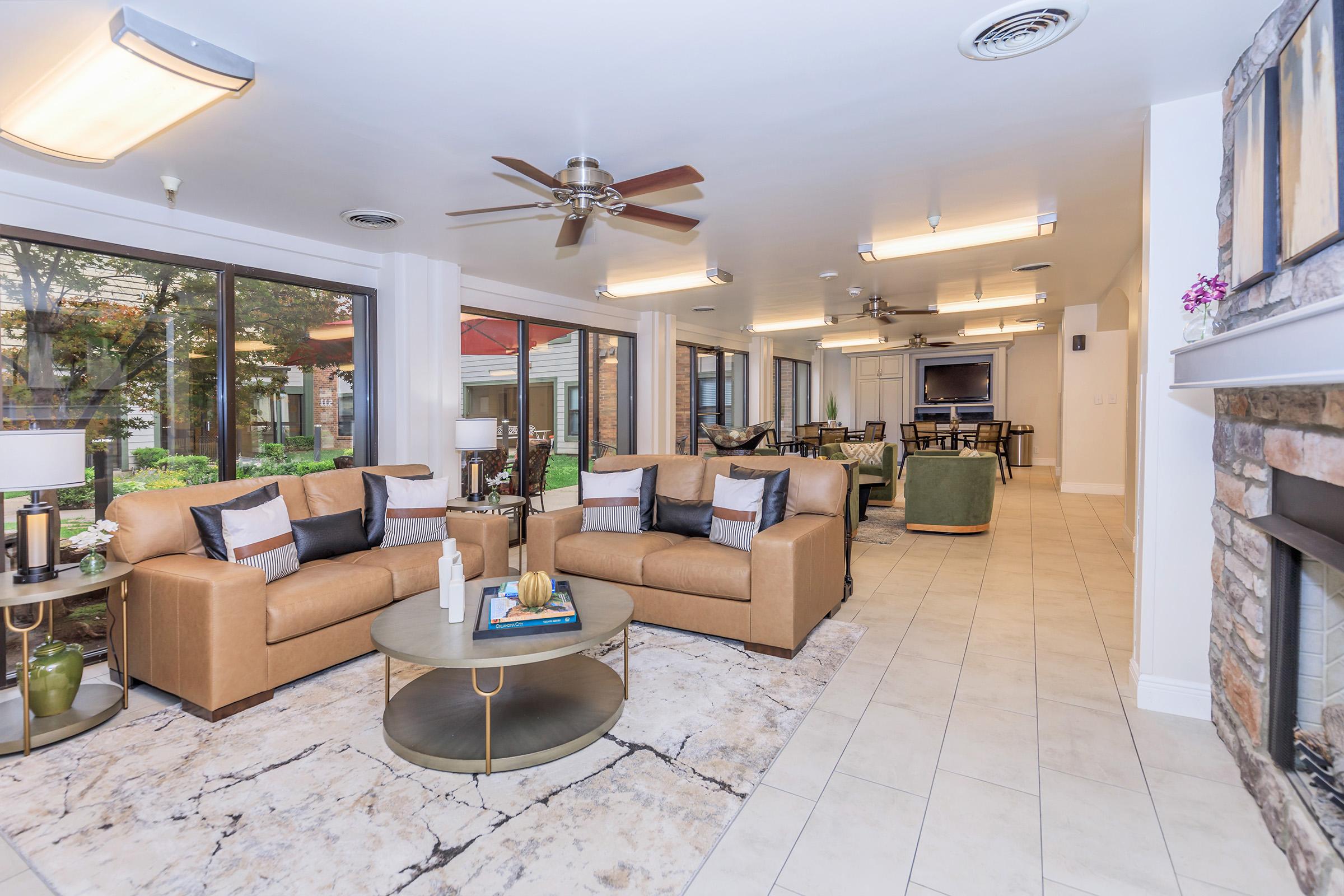 A modern living room featuring two tan leather sofas with decorative pillows, a round coffee table on a light-colored rug, and large windows letting in natural light. The space has a cozy atmosphere with potted plants, ceiling fans, and a stone fireplace, leading to an outdoor area visible through the windows.