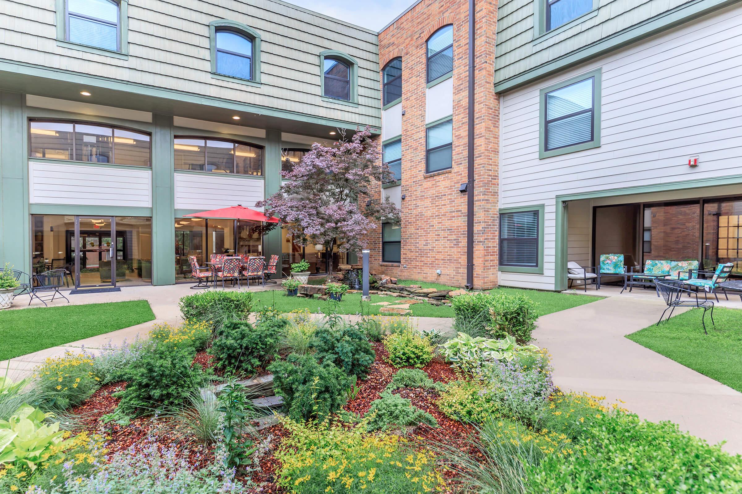 A tranquil courtyard featuring lush greenery, flower beds, and a small water feature. Surrounding the courtyard are two-story buildings with a mix of brick and siding, including outdoor seating areas and dining tables under a red umbrella. The space invites relaxation and social gatherings.