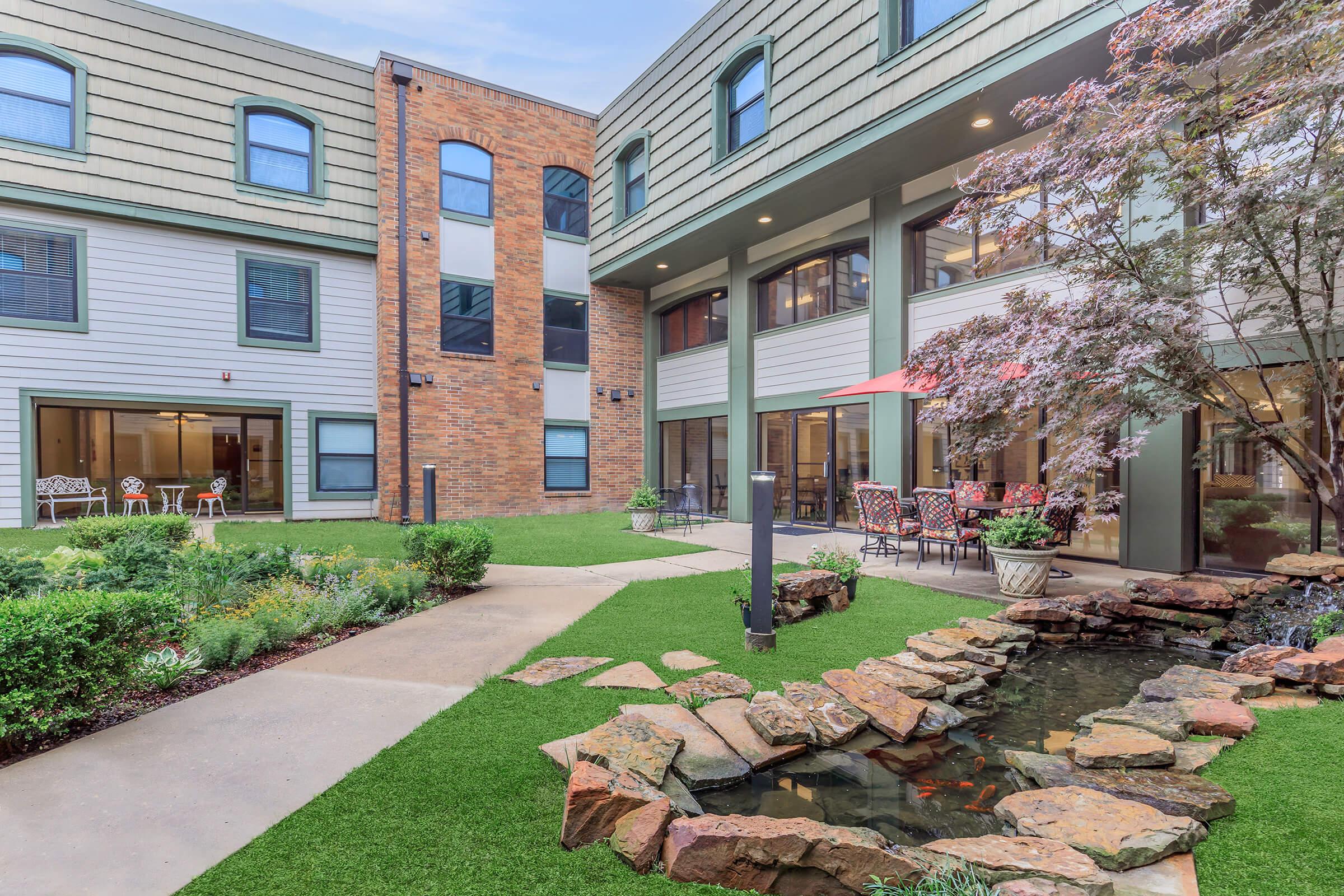 A serene courtyard featuring manicured grass, a small pond with stones, and outdoor seating areas. The space includes modern buildings with large windows, vibrant landscaping, and patio umbrellas, creating a relaxing atmosphere for gathering and enjoying nature.