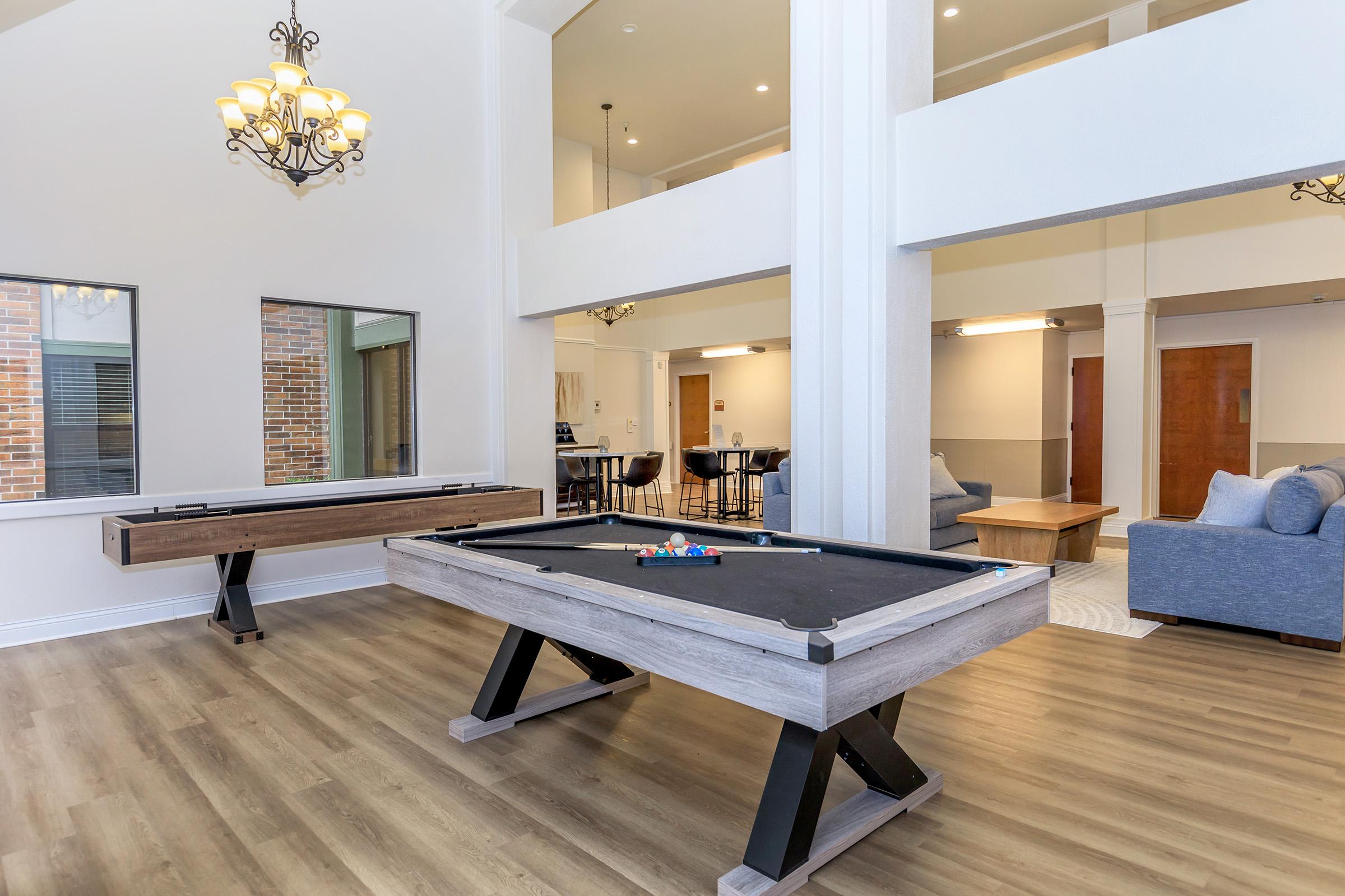 A spacious community room featuring a modern pool table in the foreground, with a decorative light fixture above. Large windows allow natural light to fill the space, which includes a cozy seating area and dining table visible in the background. The walls are painted in neutral tones, enhancing the inviting atmosphere.