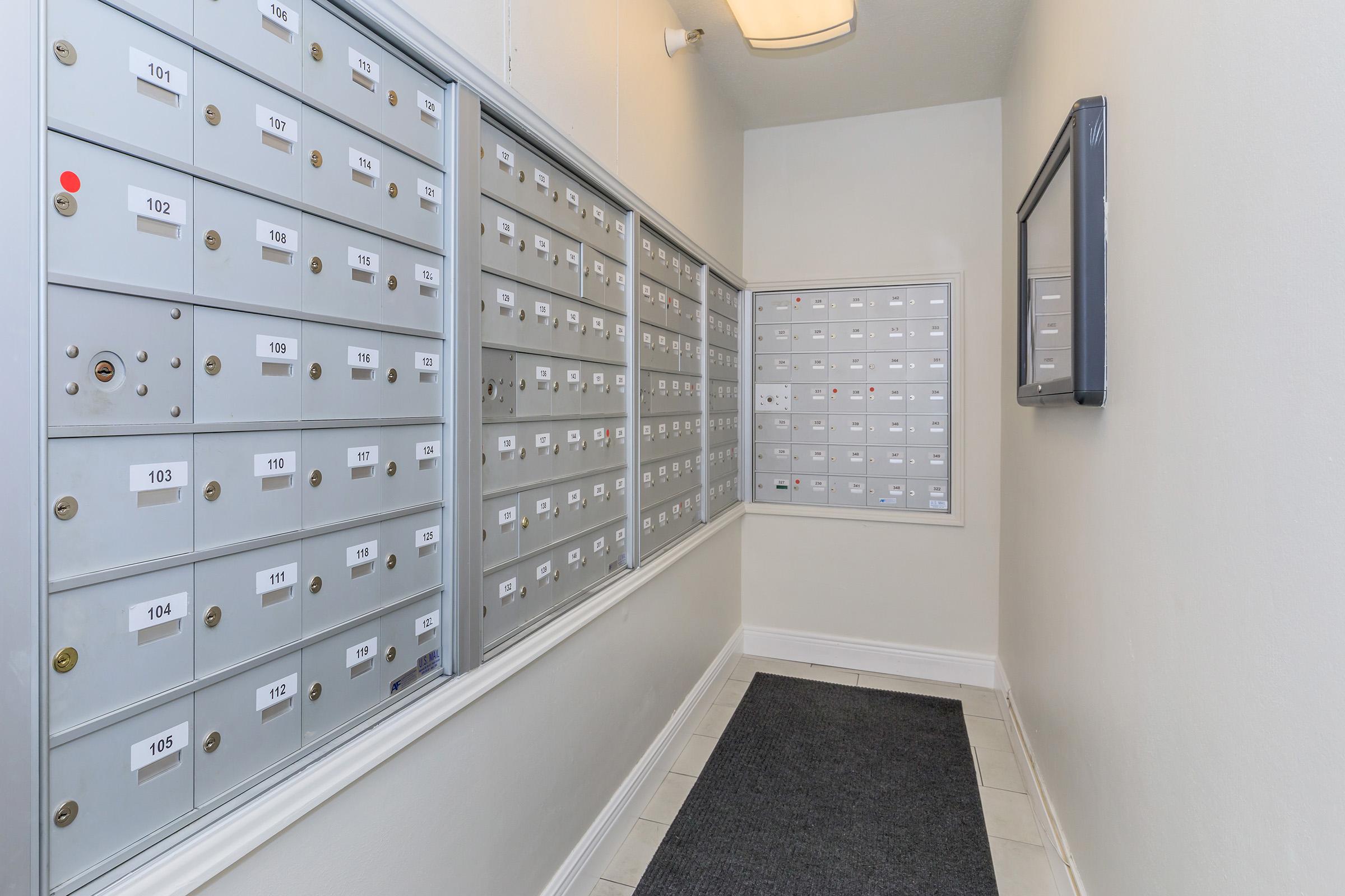 A corridor featuring multiple mailboxes on the left and a bulletin board on the right. The mailboxes are grey with numbered slots, and the wall is painted a light color. A doormat is positioned at the entrance, creating a tidy and organized appearance.