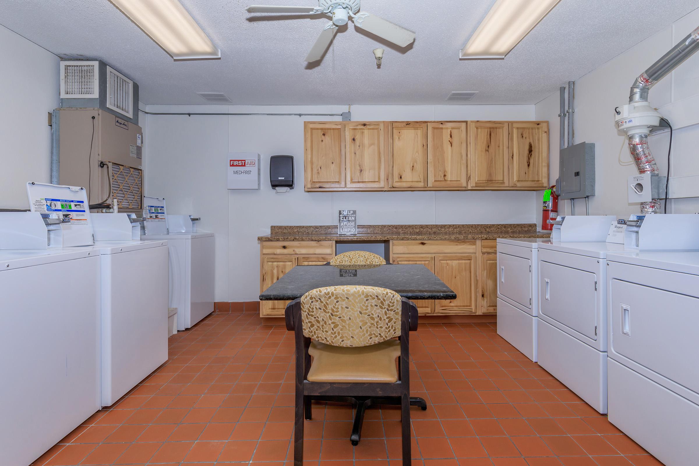 A clean and well-lit laundry room featuring several white washing machines and dryers against one wall. There is a table and a chair in the center, and wooden cabinets above the countertop provide storage. A ceiling fan is visible, and the floor is tiled in warm orange hues.