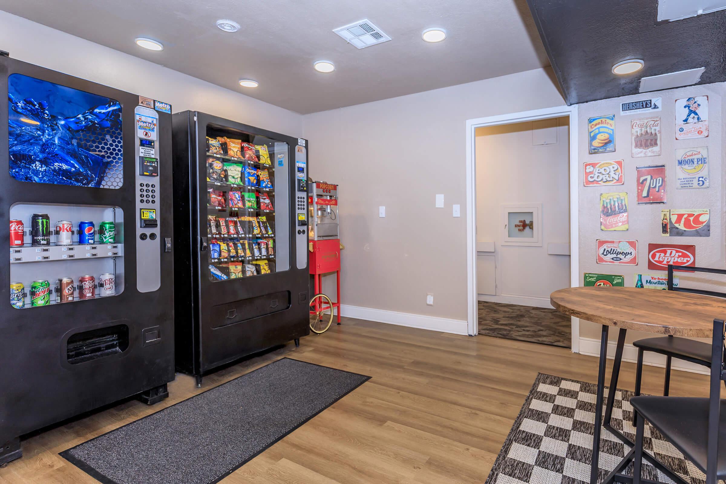 A room featuring two vending machines stocked with snacks and beverages, a small dining area with a round table and chairs, and a wall displaying various snack and beverage advertisements. The flooring combines wood and a checkered design for a modern look.