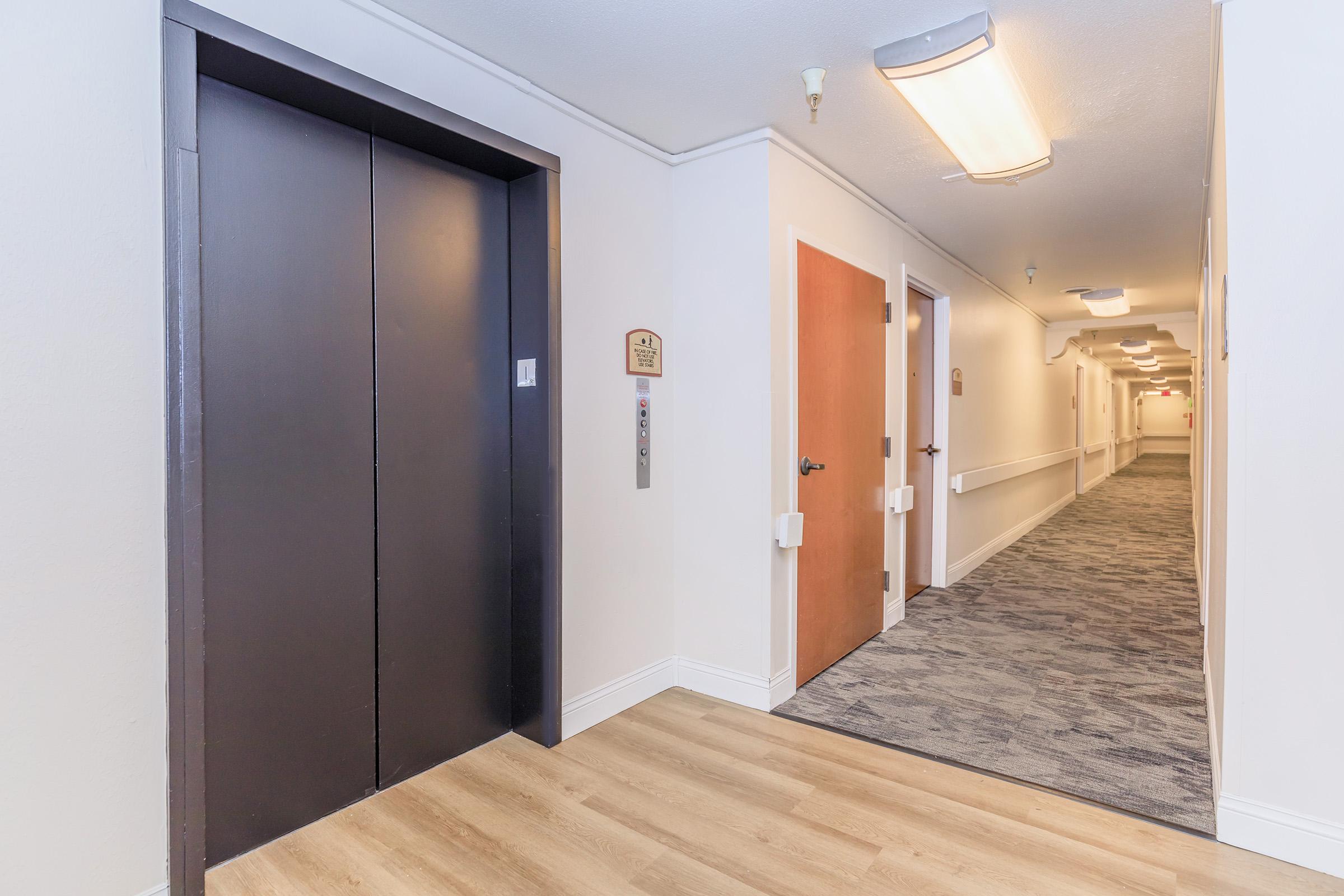 View of a hotel hallway featuring an elevator door on the left side and a closed wooden door on the right. The corridor is well-lit with ceiling lights, has beige walls, and carpeted flooring, leading further down with multiple doors along the way.