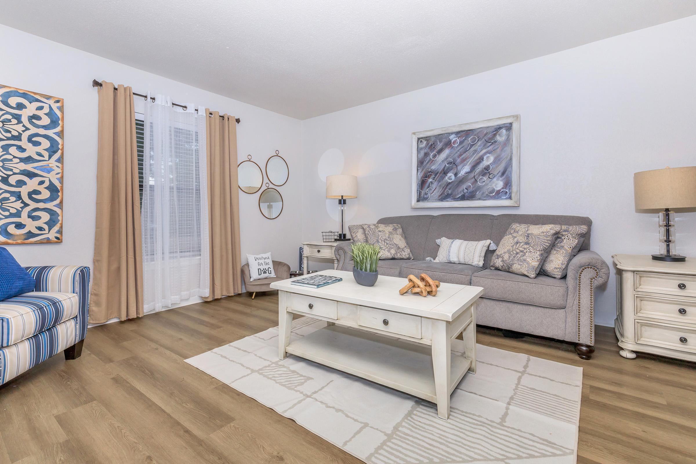 A cozy living room featuring a gray sofa with decorative pillows, an upholstered armchair with blue and white stripes, a coffee table with a decorative centerpiece, and a light-colored area rug. The walls are adorned with art and mirrors, and natural light filters in through sheer curtains.