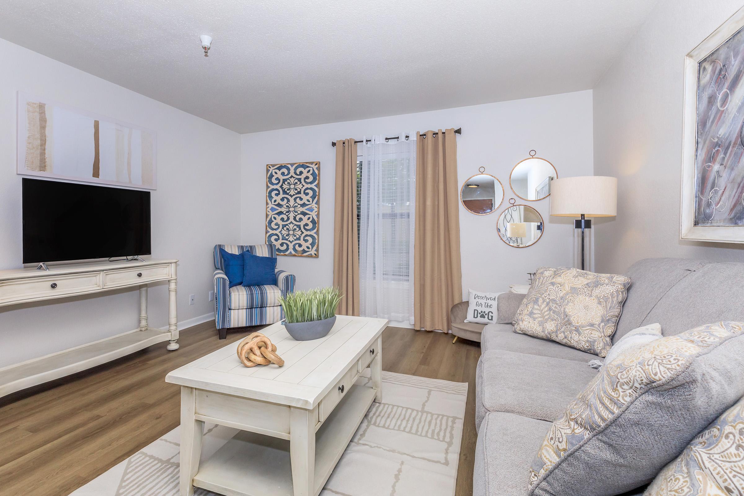 A cozy living room featuring a gray sofa with decorative cushions, a wooden coffee table, and a TV on a white console. A blue and white patterned chair sits nearby, with curtains framing a window. Decorative mirrors and wall art add style, along with a plant centerpiece on the coffee table.