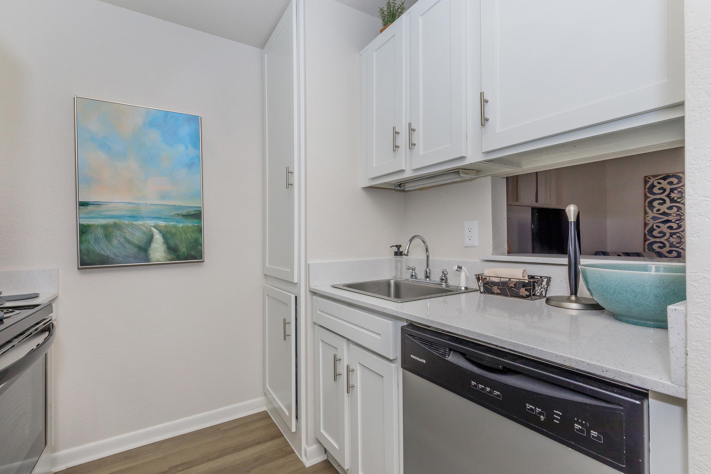 A modern kitchen with white cabinetry, a sink under a window, and appliances including a dishwasher and stove. A piece of abstract art featuring soft blue and green hues hangs on the wall. The countertop is light-colored, creating a bright and airy atmosphere.
