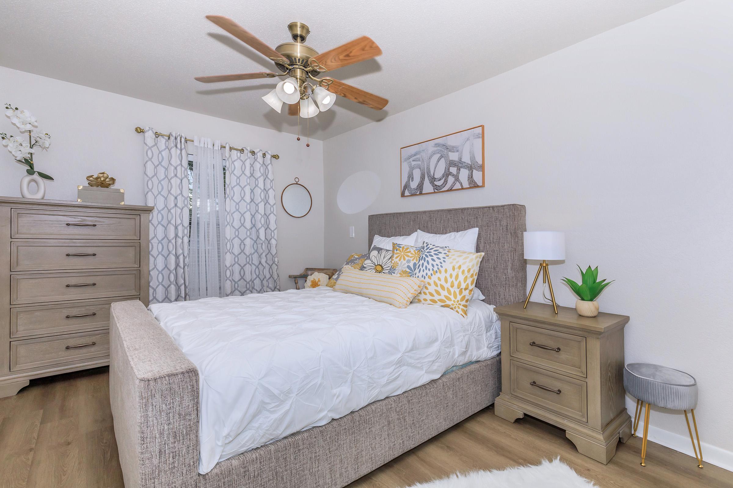 A cozy bedroom featuring a gray upholstered bed with decorative pillows in yellow and white. There is a wooden dresser, a nightstand with a lamp and small plant, and a ceiling fan. The walls are light-colored, with elegant curtains and a decorative framed picture. A soft rug is placed on the floor.