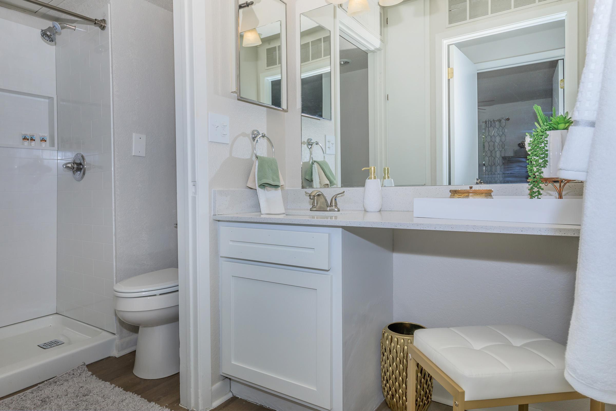 A modern bathroom featuring a white shower stall, a sleek vanity with a faucet, and a small white stool. Mirrors are mounted above the sink area, and there's a decorative plant on the countertop. The overall design is bright and airy, with light-colored walls and wood-like flooring.