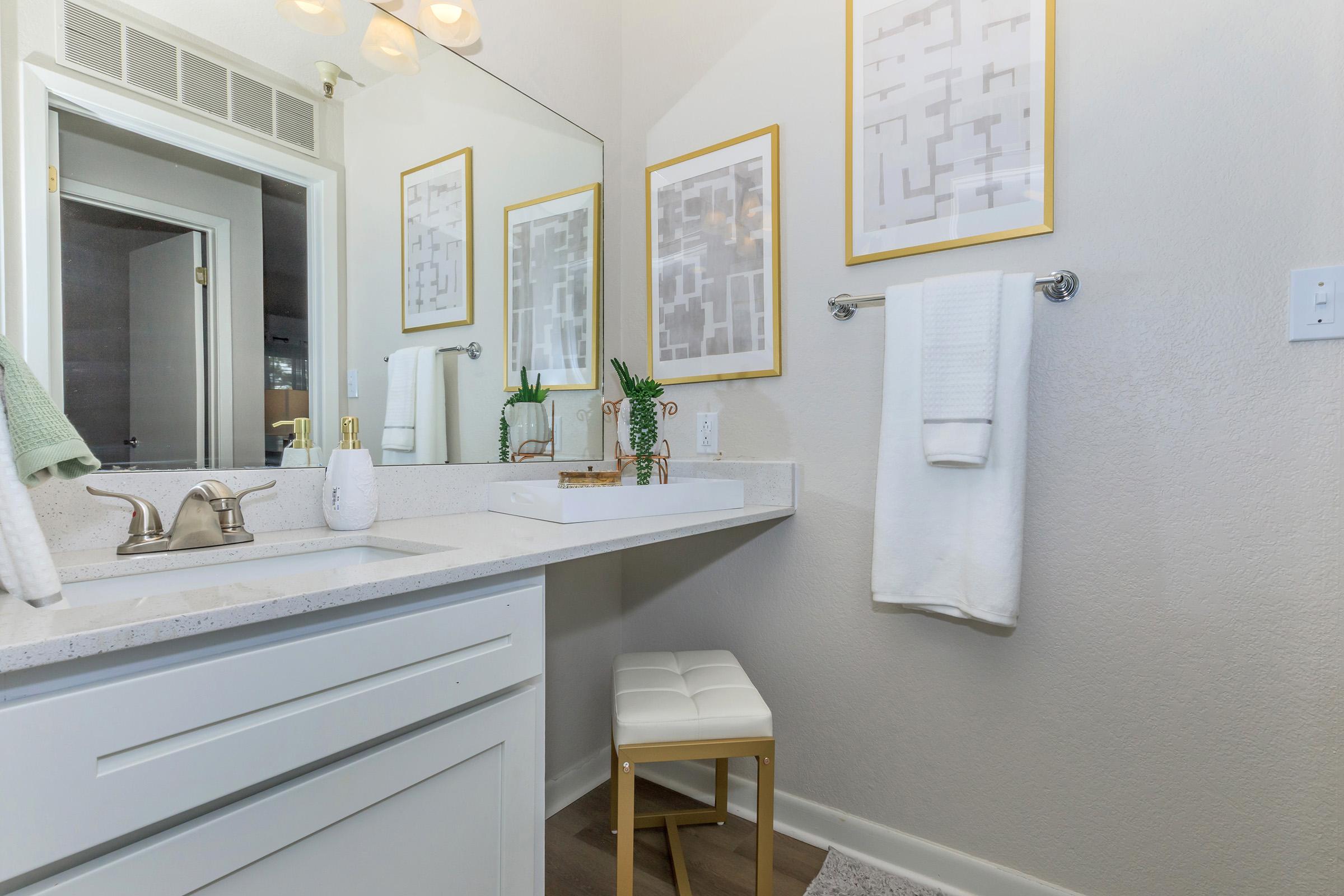 A modern bathroom featuring a white countertop with a sink, a decorative bowl, and a small plant. Above the sink are two framed abstract art prints. A towel rack holds a white towel and a light green towel. There’s a white stool nearby, and the walls are painted in a light, neutral color.