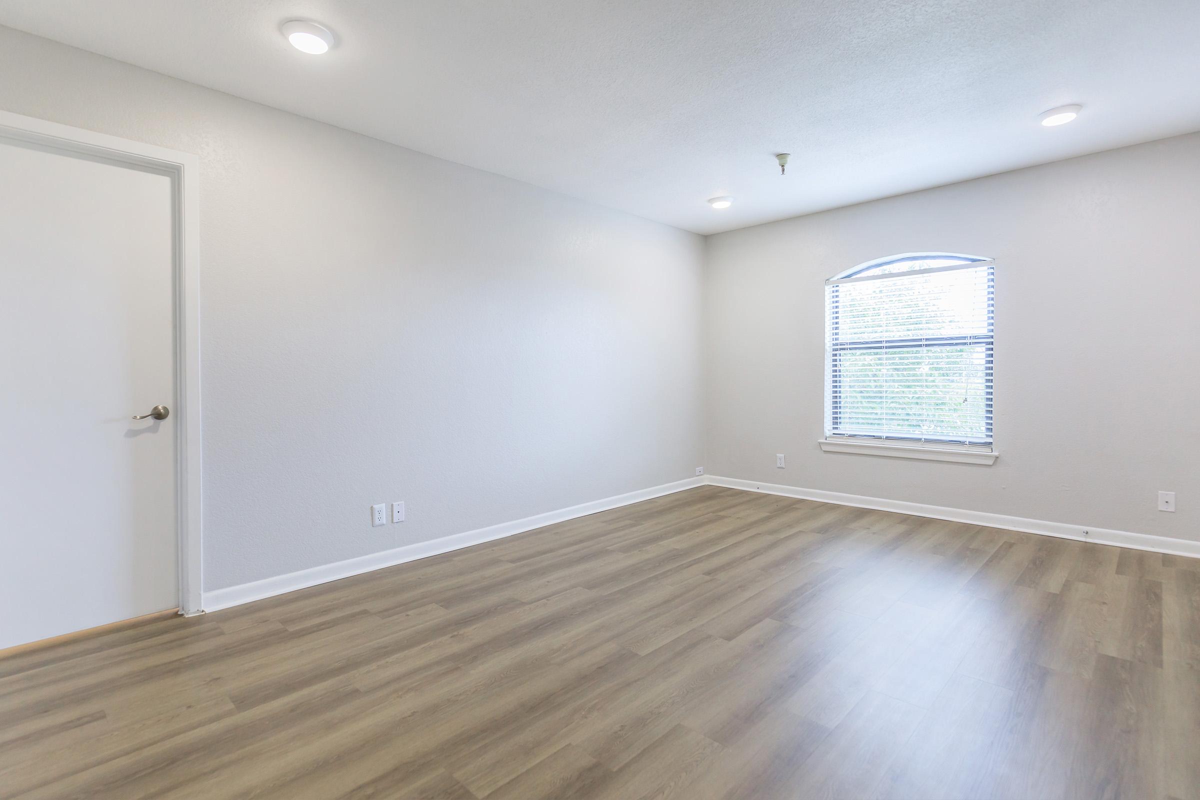A spacious, empty room featuring light gray walls and a window with white blinds. The floor is made of light-colored hardwood. There is a white door on the left, and the room appears well-lit with ceiling lights. The atmosphere is clean and ready for furnishing.