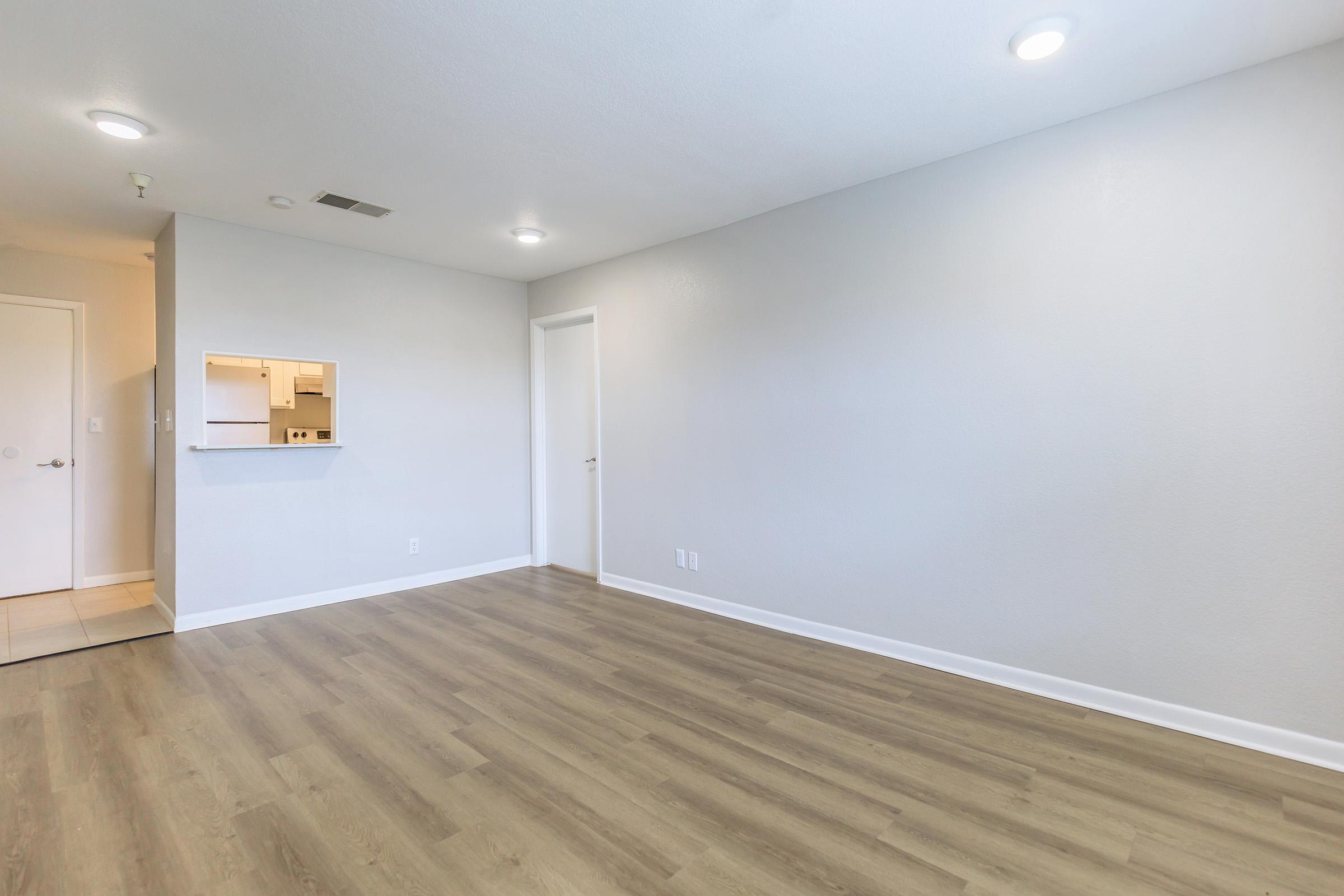 A spacious, empty room with light gray walls and a wood-style floor. There is a small opening to a kitchen area on one side, and a door on the opposite side leads to another room. The room is well-lit with recessed lighting in the ceiling, creating a bright and inviting atmosphere.