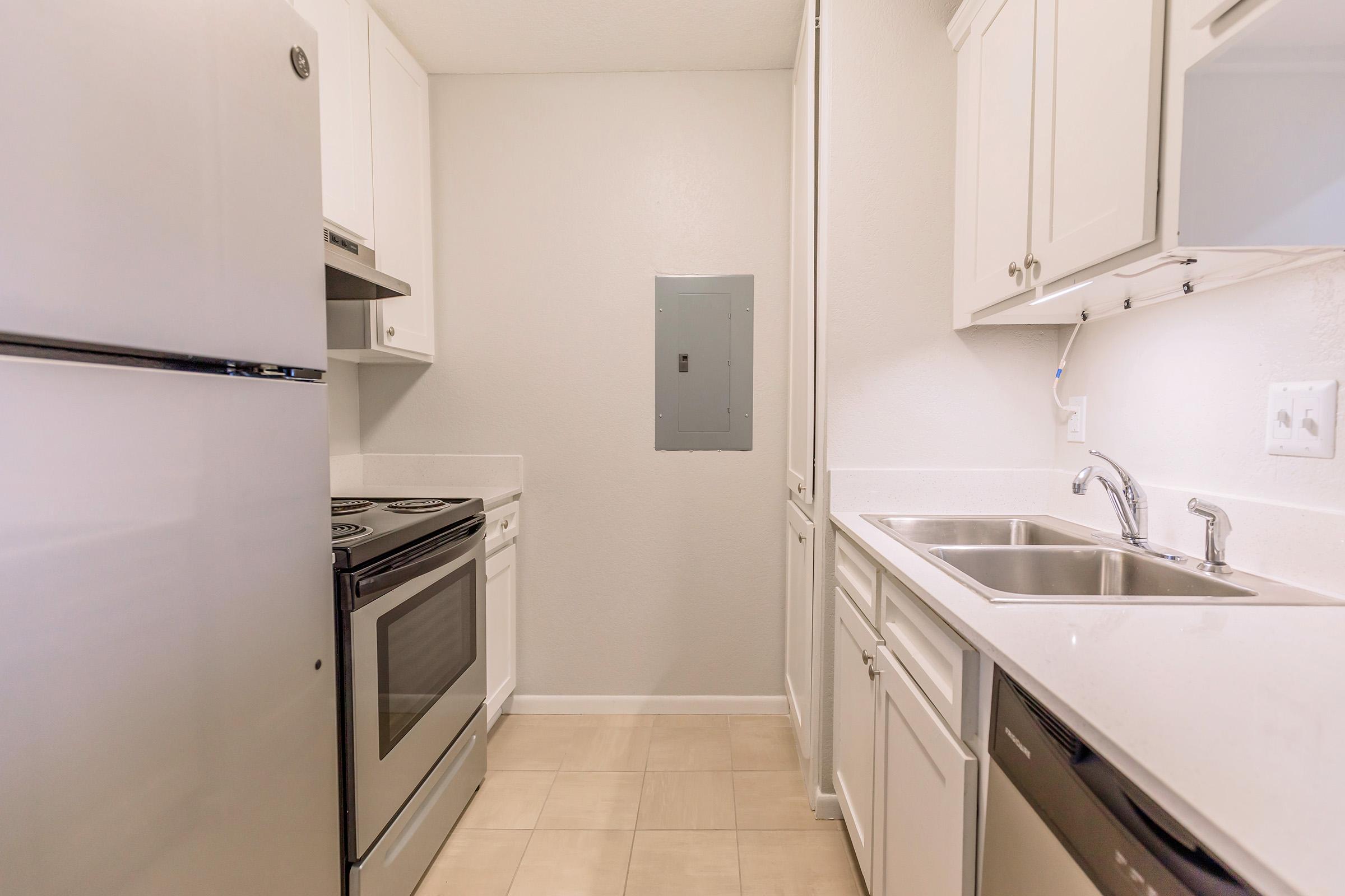 A small kitchen featuring white cabinetry, a silver refrigerator, an oven with a stovetop, and a double sink. The walls are painted light gray, and the floor has beige tiles. A power panel is visible on one wall. The overall layout is functional and compact, suitable for a small living space.