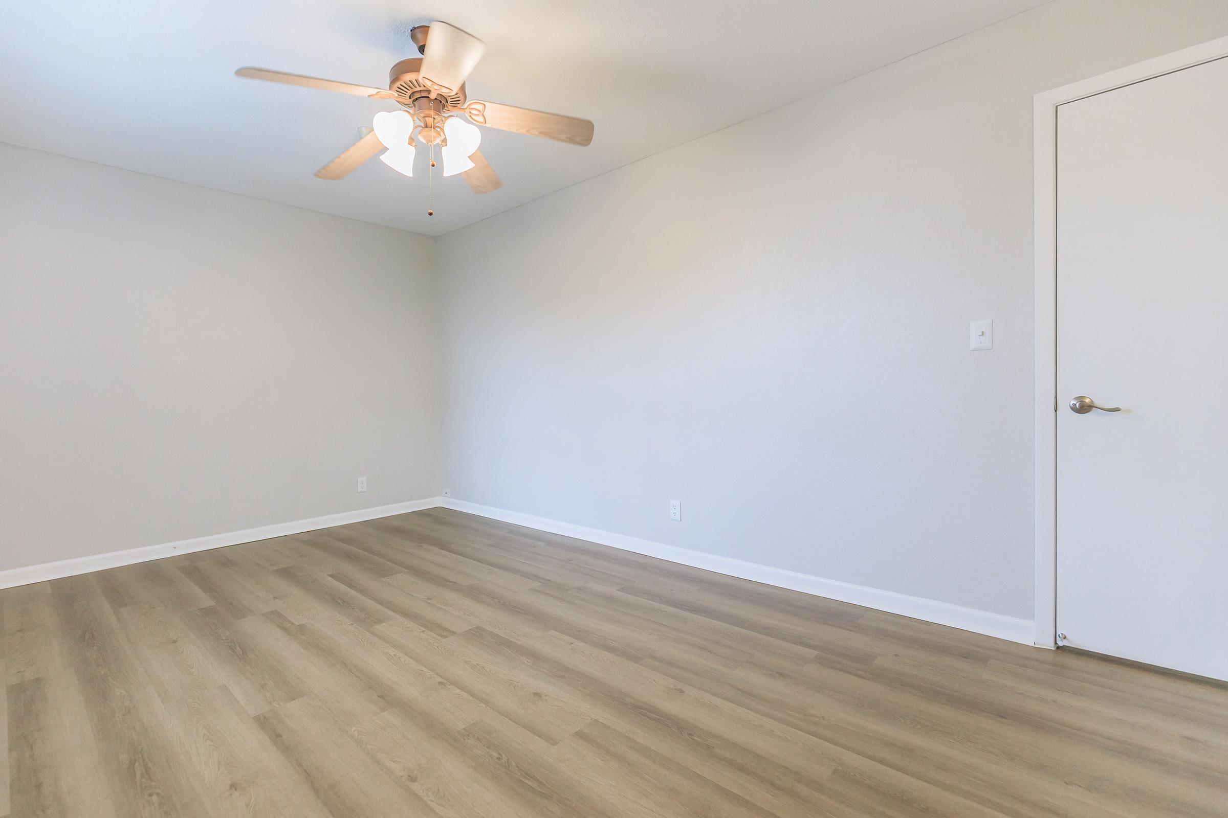Empty room with light gray walls and a wooden floor. A ceiling fan with wooden blades is centered on the ceiling. A white door is visible on the right side of the image. The space is well-lit and appears to be freshly painted and clean.