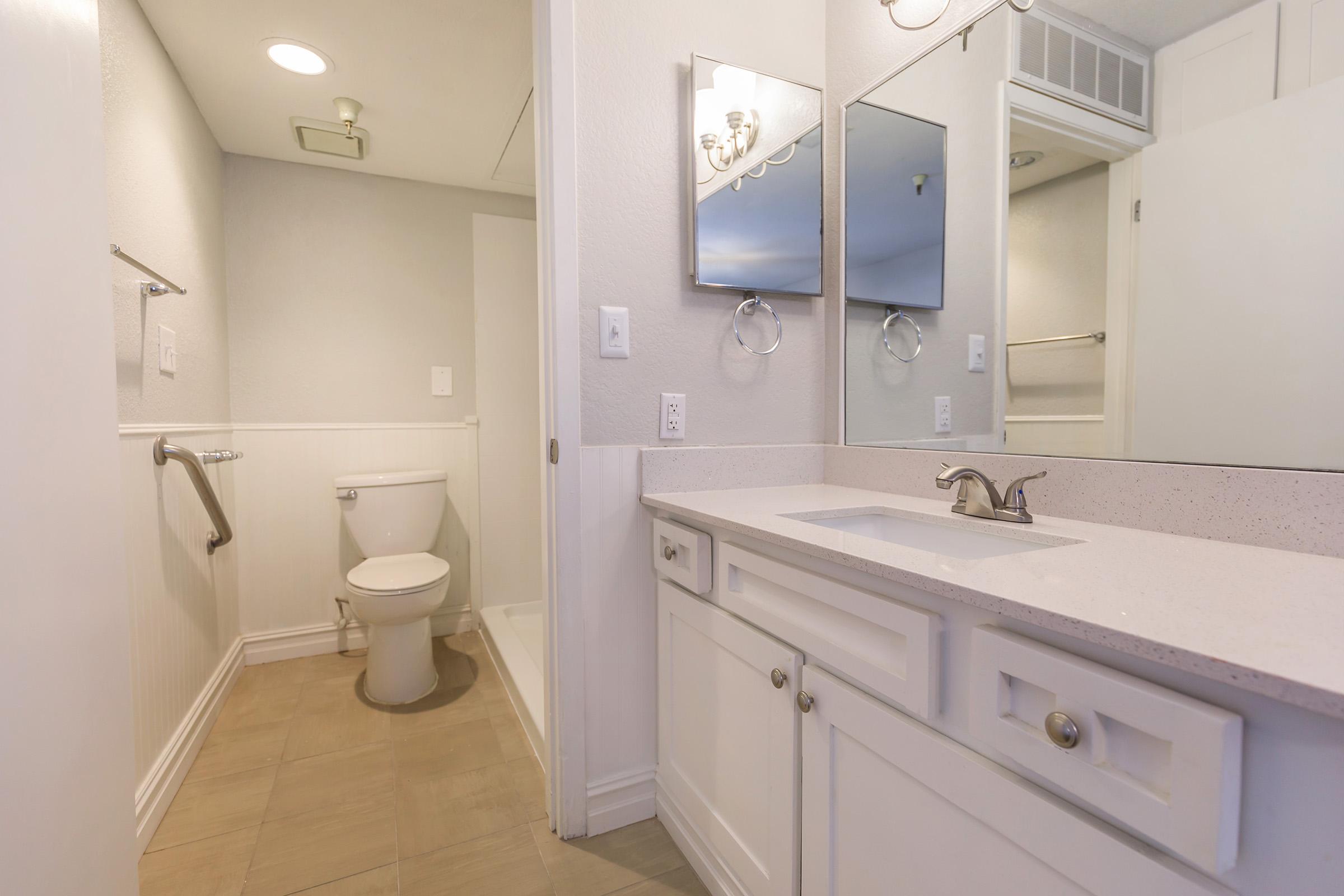 A clean, modern bathroom featuring a white vanity with a sink, a toilet, and two mirrors above the sink. The walls are painted in light colors, and the floor is tiled. There is a shower area visible, along with a grab bar for safety. The overall design is simple and well-lit.