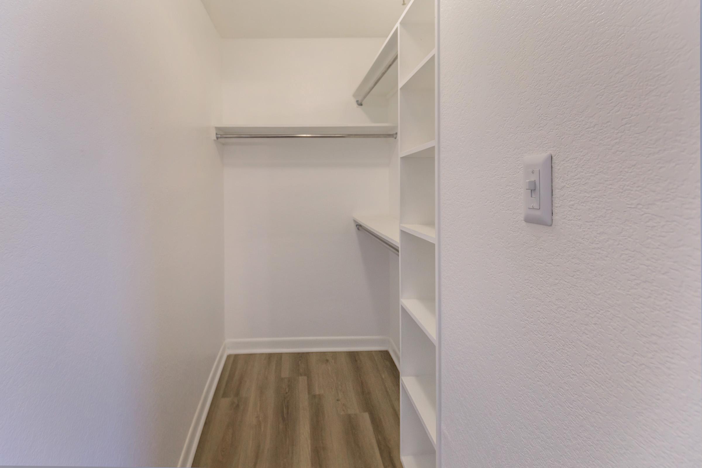 A spacious walk-in closet featuring white walls, a wooden floor, and built-in shelving. The closet includes a hanging rod along one side and shelves on the other, providing ample storage space for clothing and accessories.