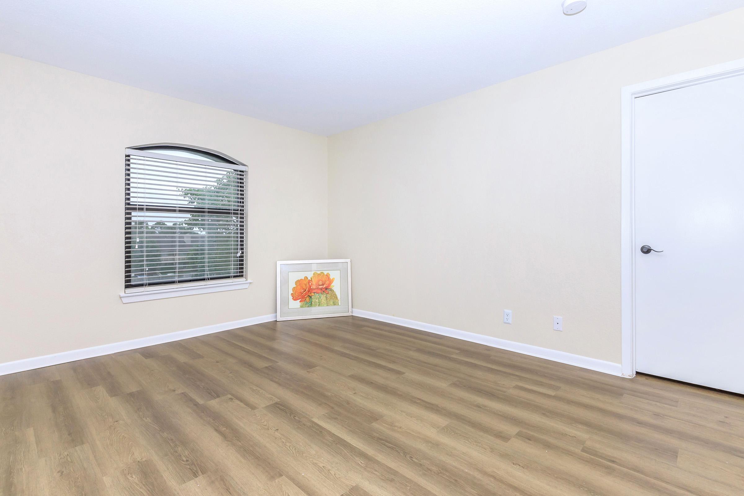 A simple, empty room with a light yellow wall and hardwood flooring. There is a window with blinds on the left side and a framed picture of flowers leaning against the wall. A closed white door is visible in the right corner. The space is bright and uncluttered.
